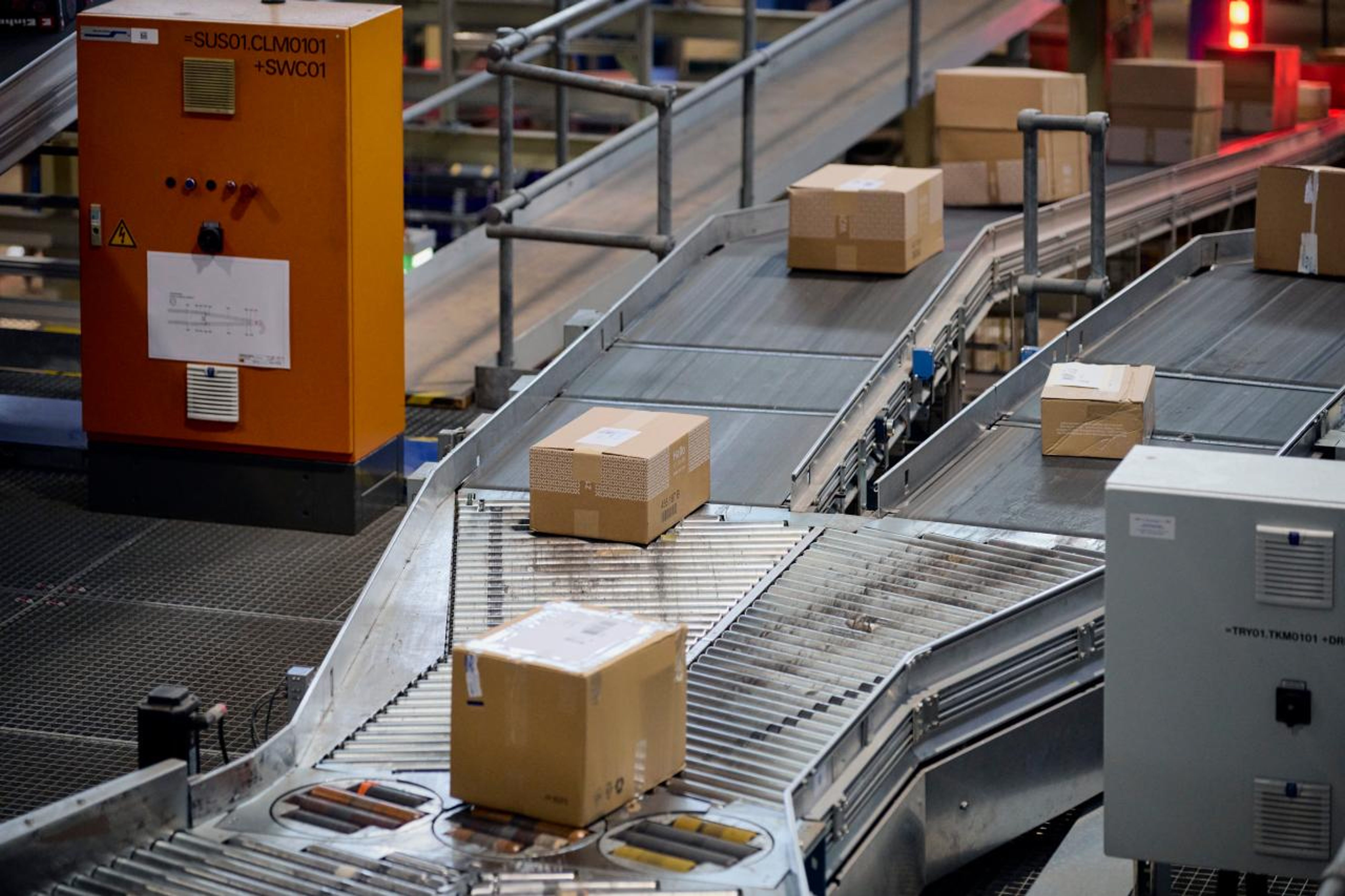 Un système de convoyage automatisé transporte plusieurs colis sur des voies de circulation dans un centre logistique. Un élément de contrôle orange est visible, ce qui indique un environnement industriel. La scène se concentre sur le mouvement efficace des envois.
