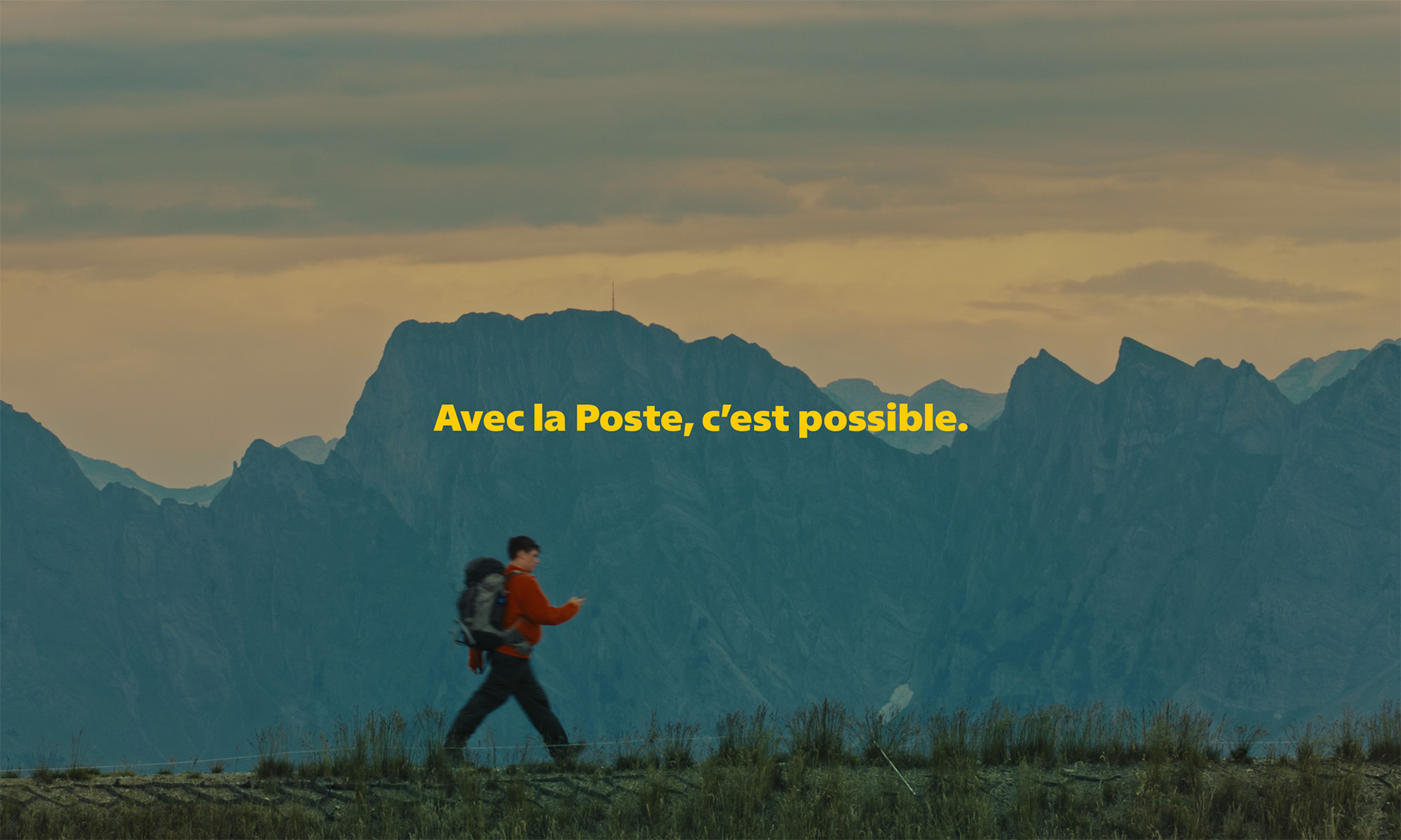 Une personne se tient devant un véhicule de transport ouvert et tient un smartphone montrant une interface jaune avec plusieurs icônes. Devant la personne se trouvent des cartons et des surfaces en bois. À l’arrière‑plan, on aperçoit des arbres, un bâtiment et une structure couverte. Le texte jaune « Die Post macht’s möglich. » apparaît au centre de l’image.