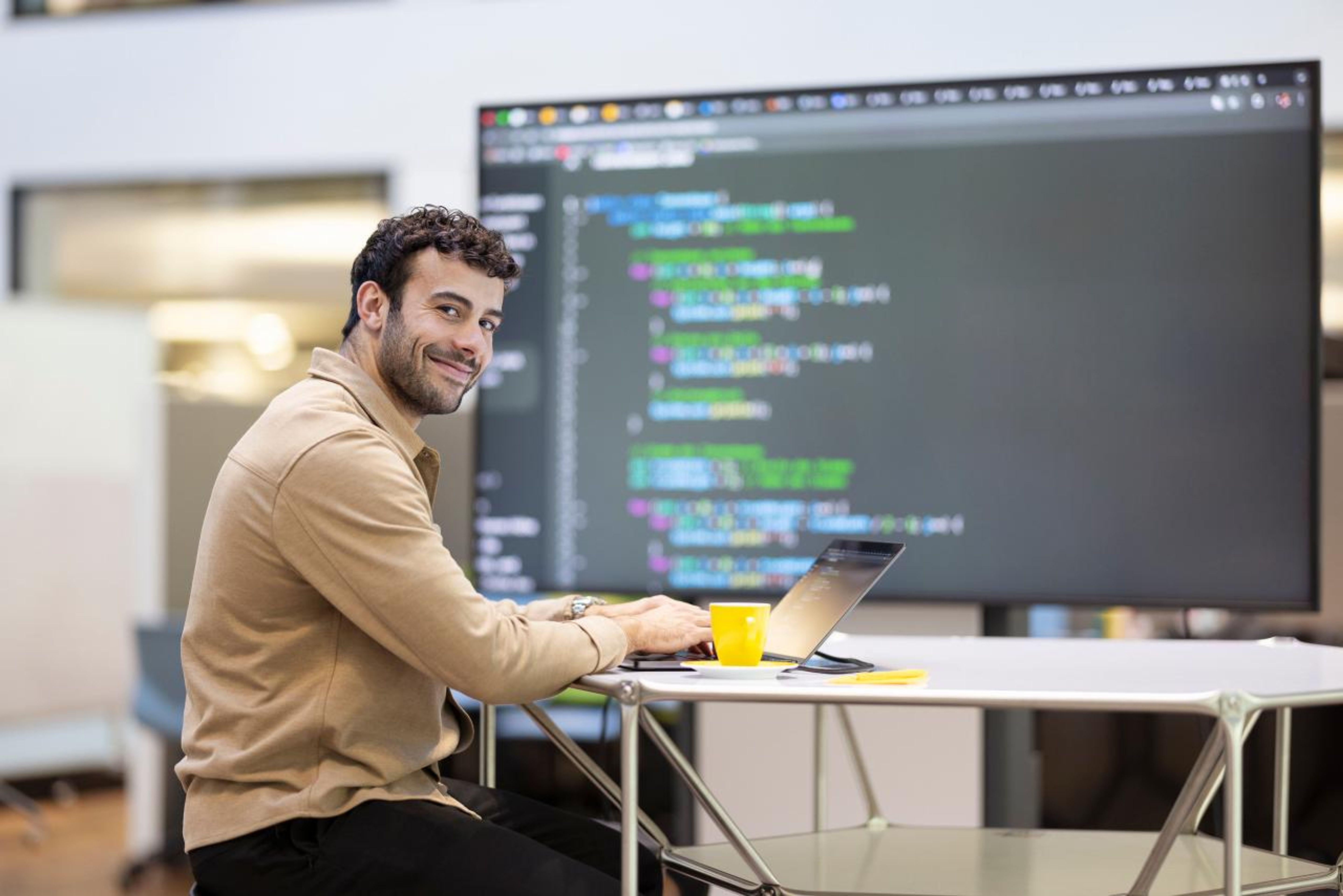 Un homme est assis à une table et travaille sur un ordinateur portable devant un grand écran sur lequel le code s’affiche. Il sourit à la caméra, l’environnement est moderne et convivial. La salle est bien éclairée et l’atmosphère est professionnelle et créative,