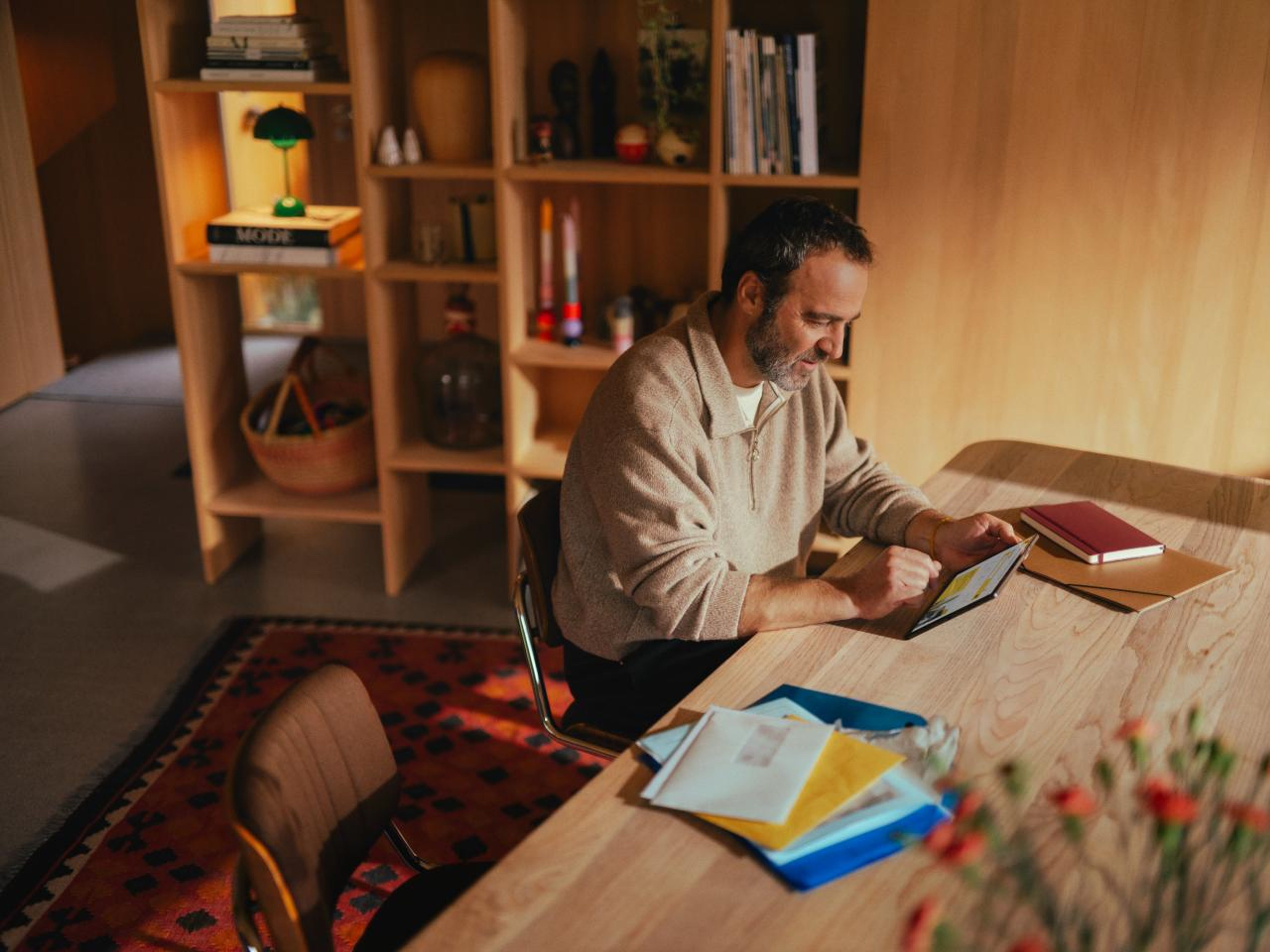 Un homme assis à une table en bois dans une pièce confortable utilise une tablette. Des lettres et un bloc-notes sont posés sur la table. L’arrière-plan montre une étagère avec des livres et des décorations, créant une atmosphère chaleureuse et accueillante.
