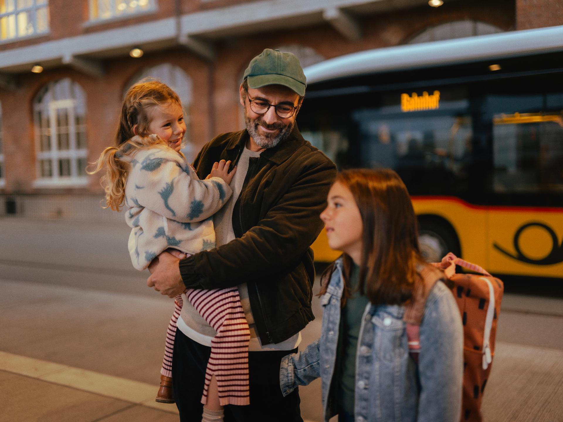 Three people are standing at a bus stop in front of a yellow bus. One person is holding a child, while another person with a backpack stands nearby. In the background, a brick building with illuminated windows is visible.