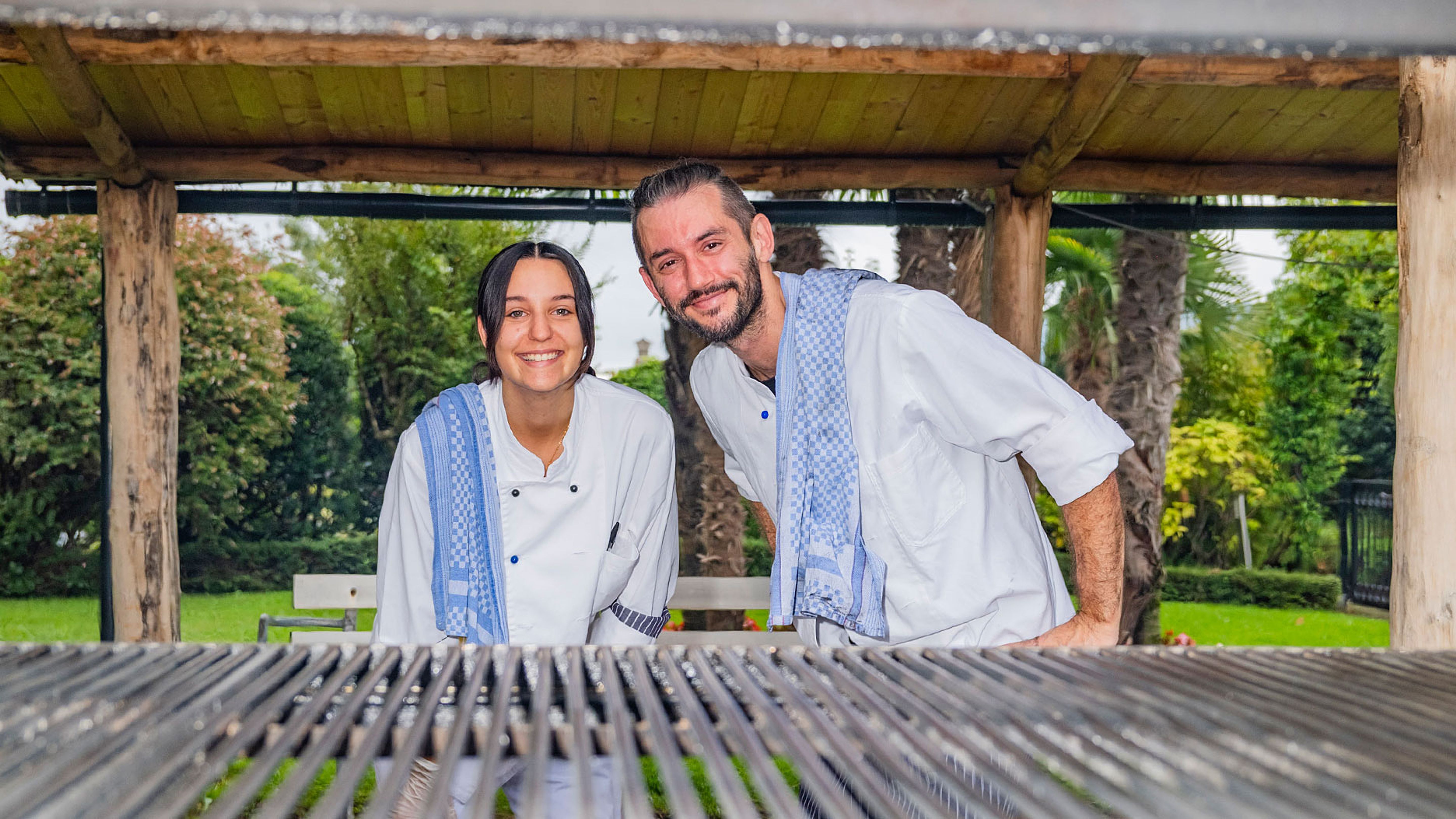 Zwei Personen in weissen Küchenjacken und blaue Tücher in einem holzüberdachten Gartenbereich mit Grillrost.