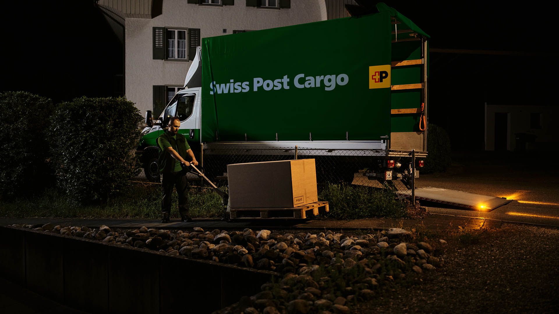 Un corriere notturno di Swiss Post Cargo consegna beni medicinali con un carrello a mano, con un veicolo di consegna sullo sfondo.