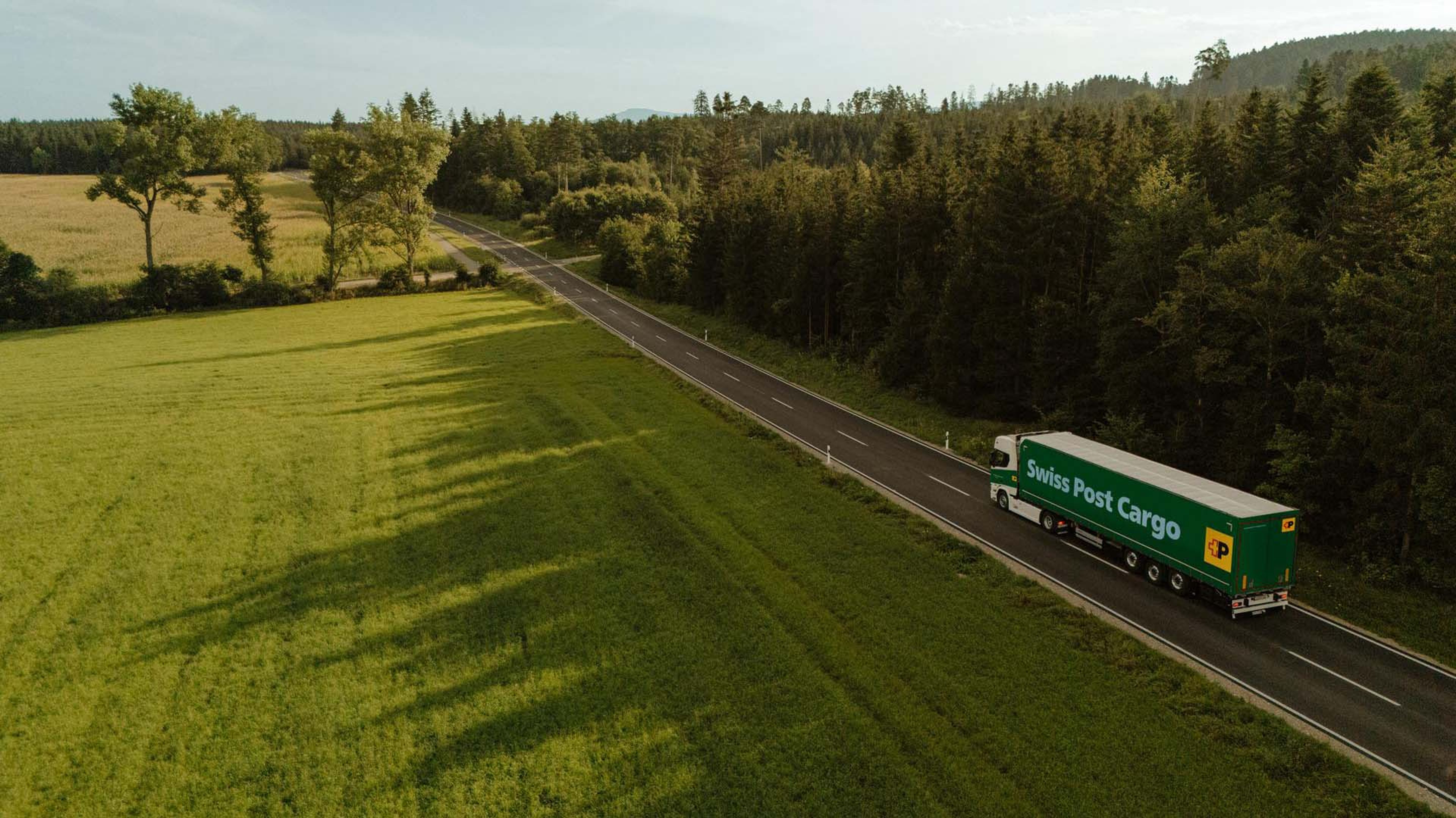 Swiss Post Cargo LKW fährt auf einer Landstrasse zwischen einer Wiese und einem Wald.