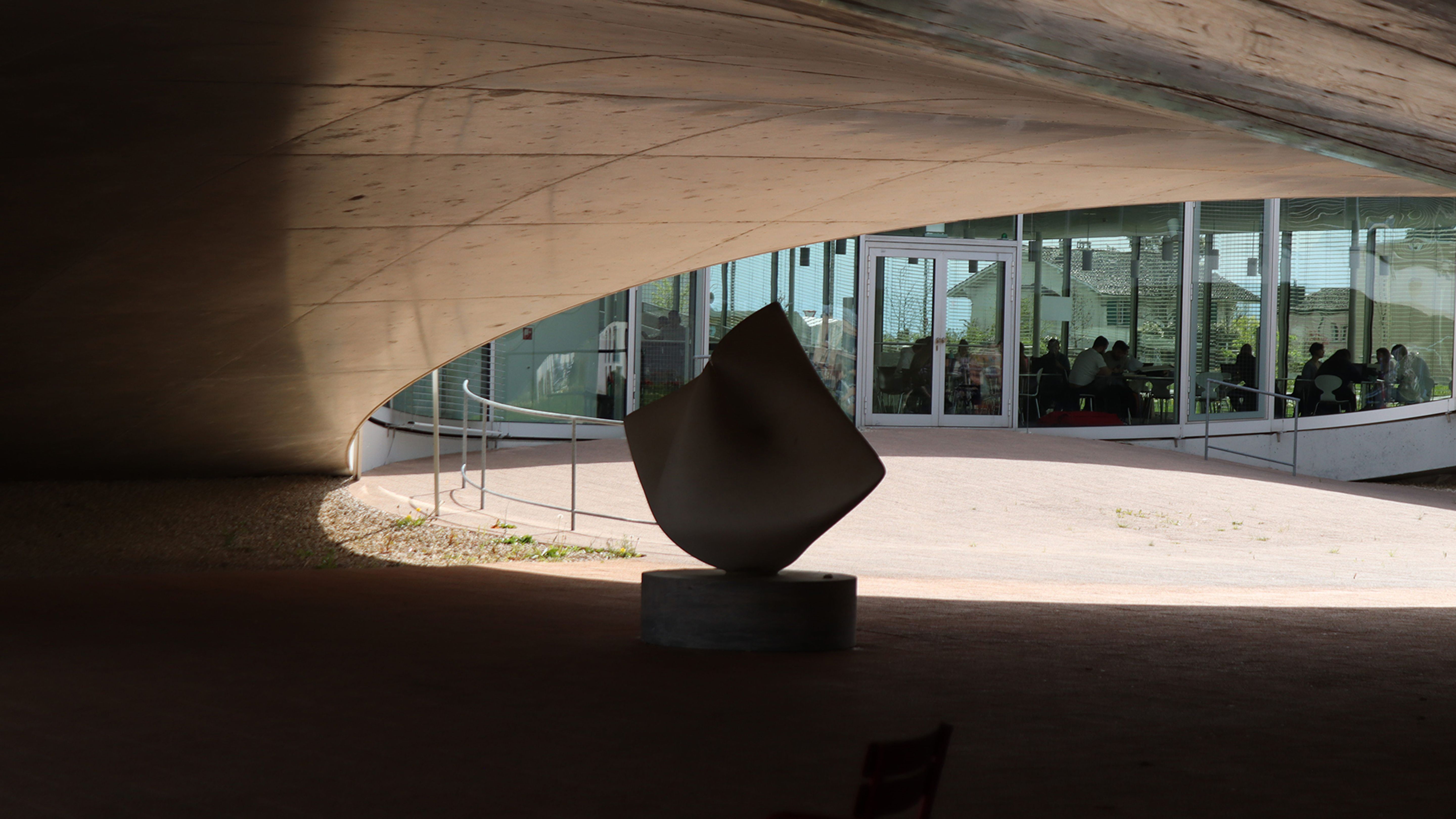 Una scultura è collocata sotto un soffitto in cemento ricurvo, con finestre in vetro e persone sedute sullo sfondo.