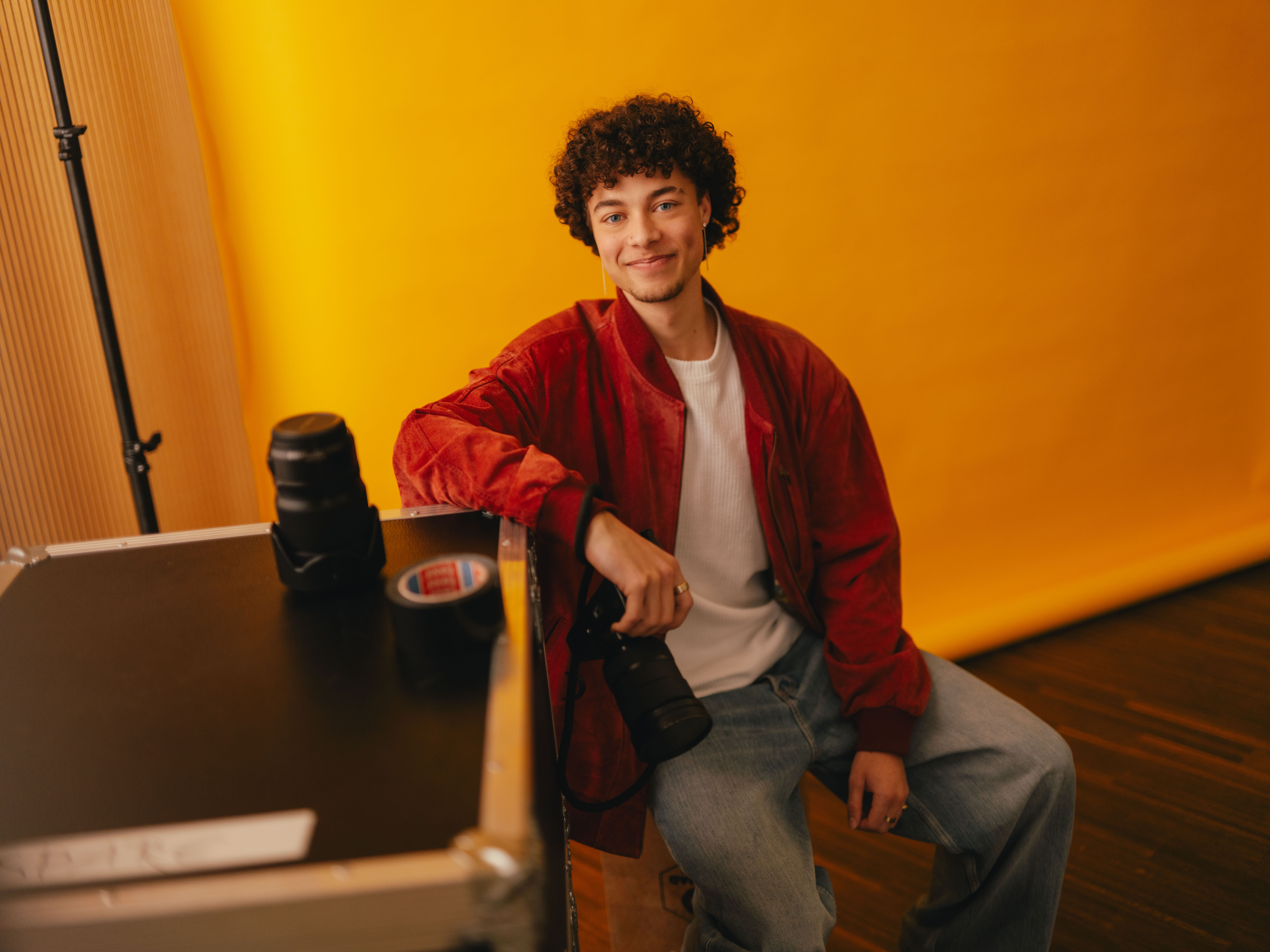 A person is sitting on a transport case against a bright yellow background. On the case are camera lenses and a roll of adhesive tape. The person is wearing a red jacket, a white T‑shirt and loose-fitting jeans and holding a camera loosely in one hand. The room looks like a photo studio with professional equipment.