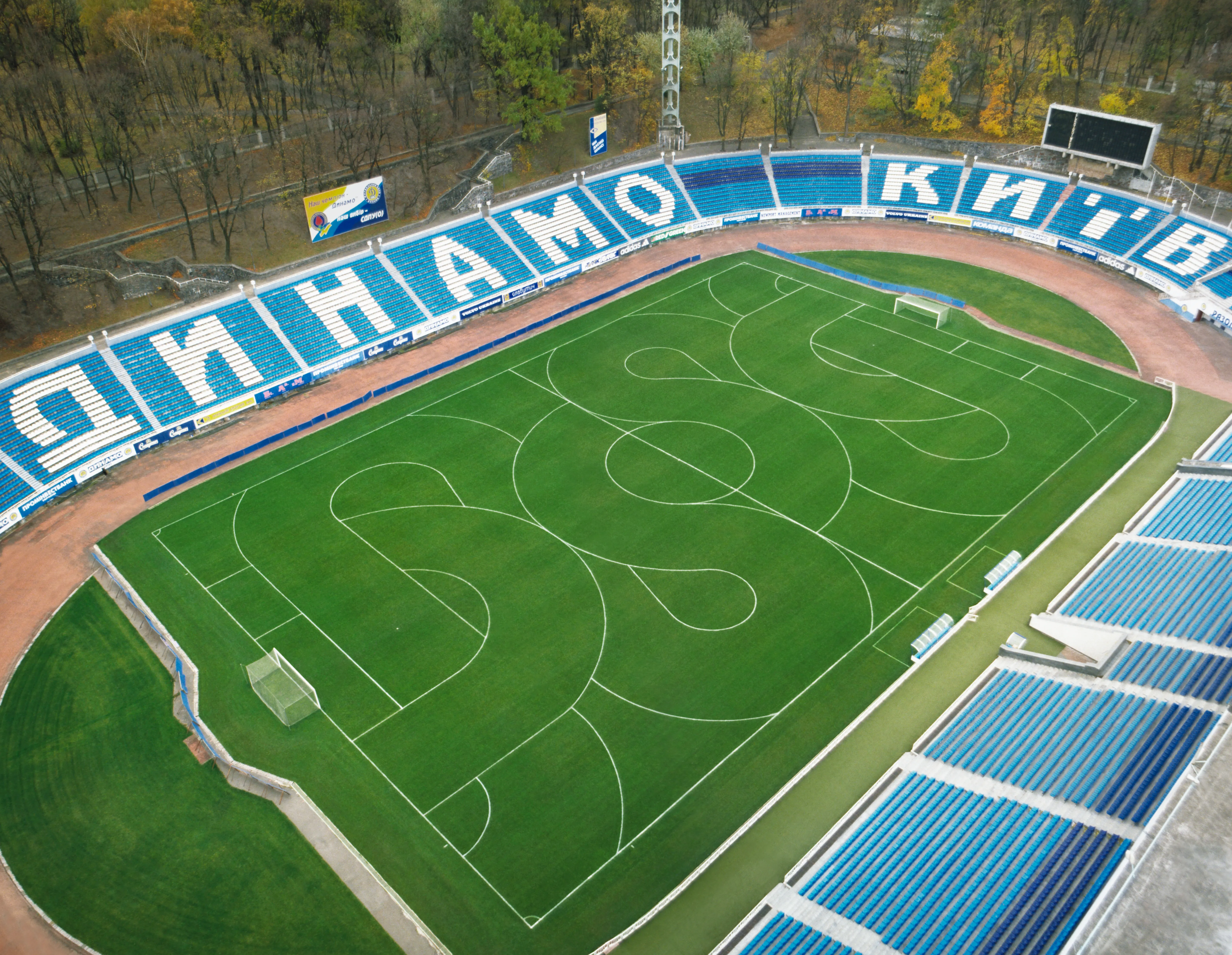 On aperçoit un stade de football avec des lignes fauchées sur le terrain, des tribunes bleues vides et la forêt environnante.
