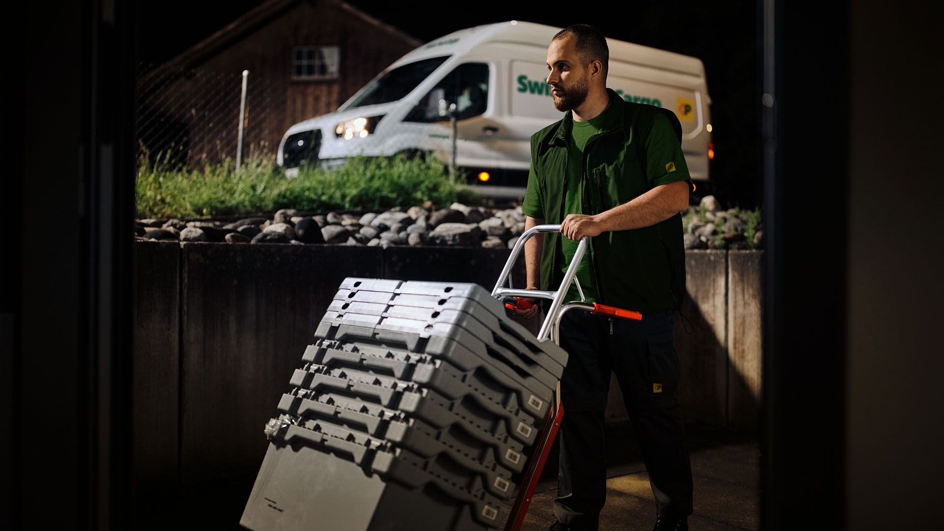 Un corriere notturno di Swiss Post Cargo consegna beni medicinali con un carrello a mano, con un veicolo di consegna sullo sfondo.