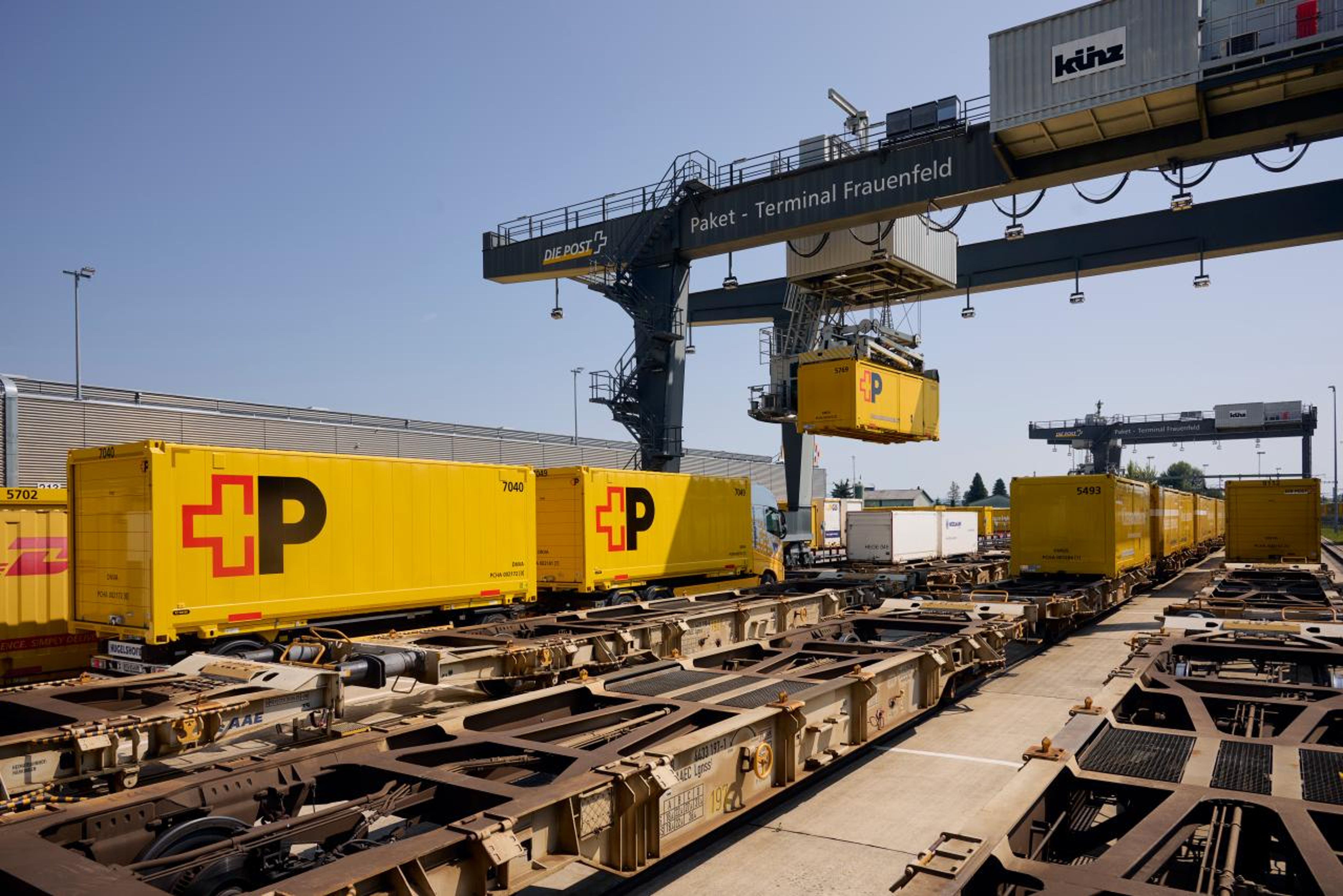 Des conteneurs postaux jaunes avec le logo de la Poste sont posés sur des wagons au terminal de colis de Frauenfeld. Une grande grue soulève un conteneur. La scène montre une logistique efficace sous un ciel clair dans un centre de transbordement moderne. L’atmosphère est animée et structurée.