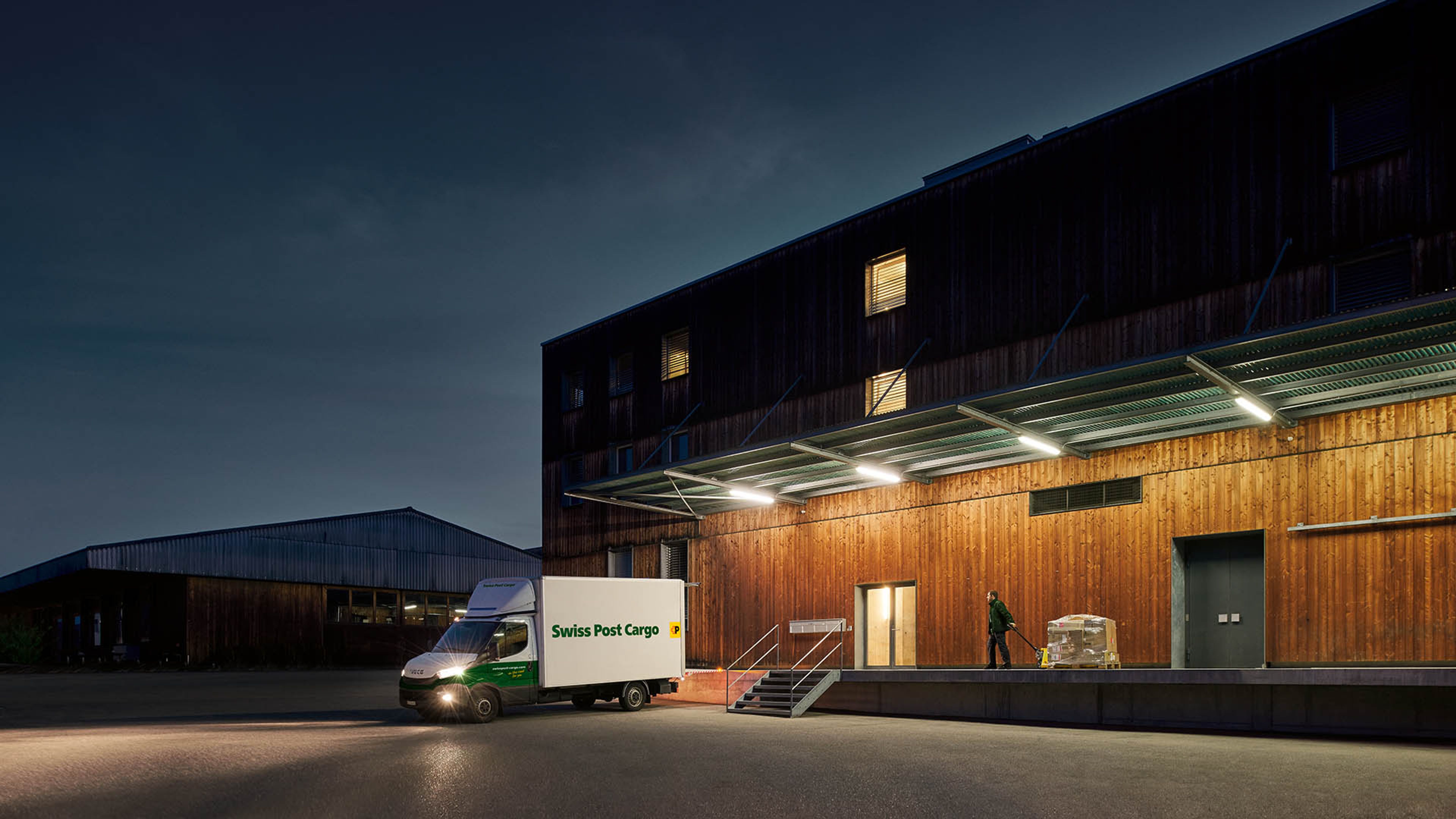 A Swiss Post Cargo night courier delivers goods to a warehouse building before dawn.