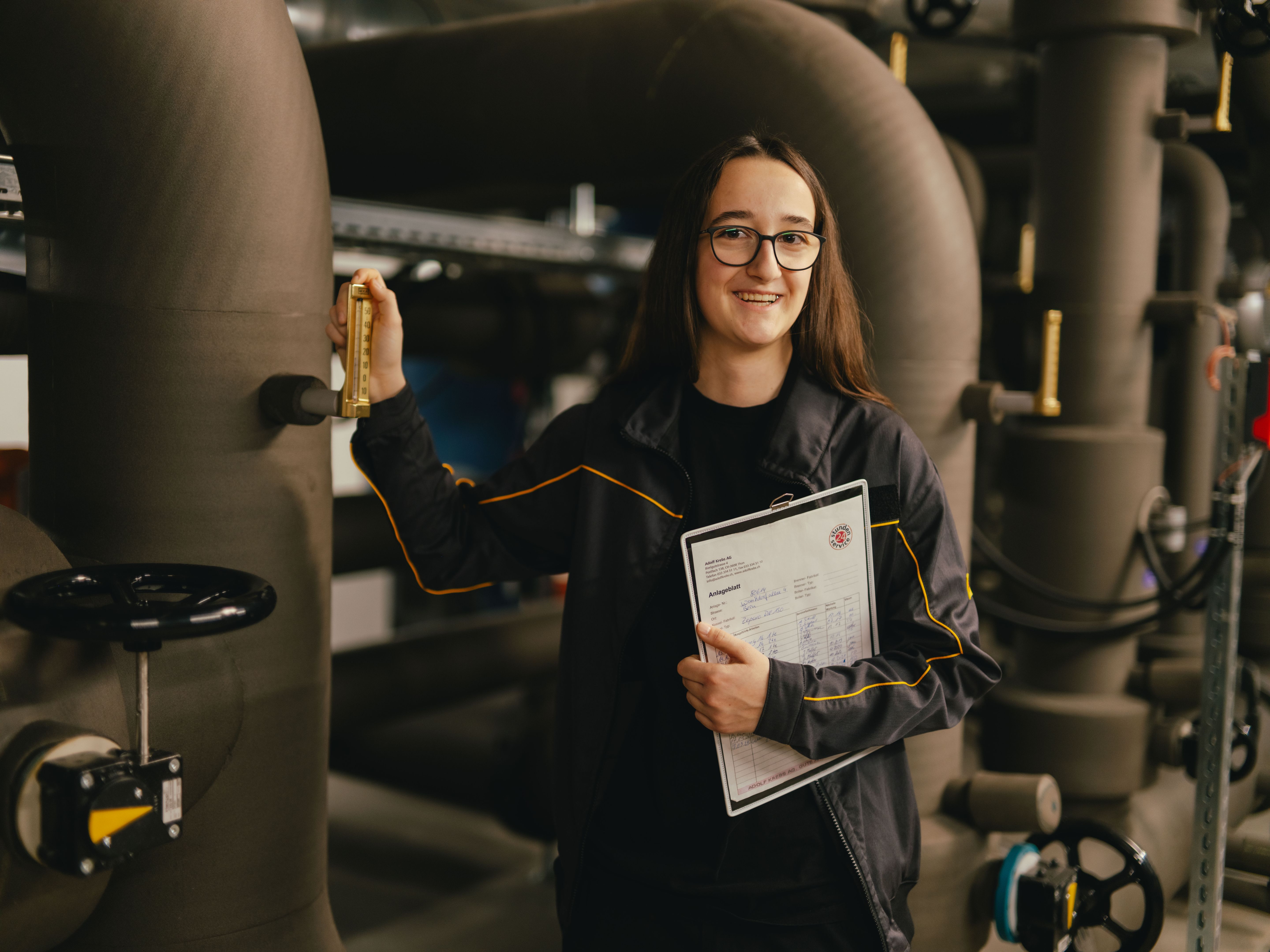 Eine Person steht in einer technischen Anlage zwischen grossen Rohrleitungen und hält ein Klemmbrett mit technischen Unterlagen. Mit der anderen Hand bedient die Person einen metallenen Hebel an einem der Rohre. Die Umgebung ist industriell mit Ventilen, Leitungen und gedämpfter Beleuchtung.