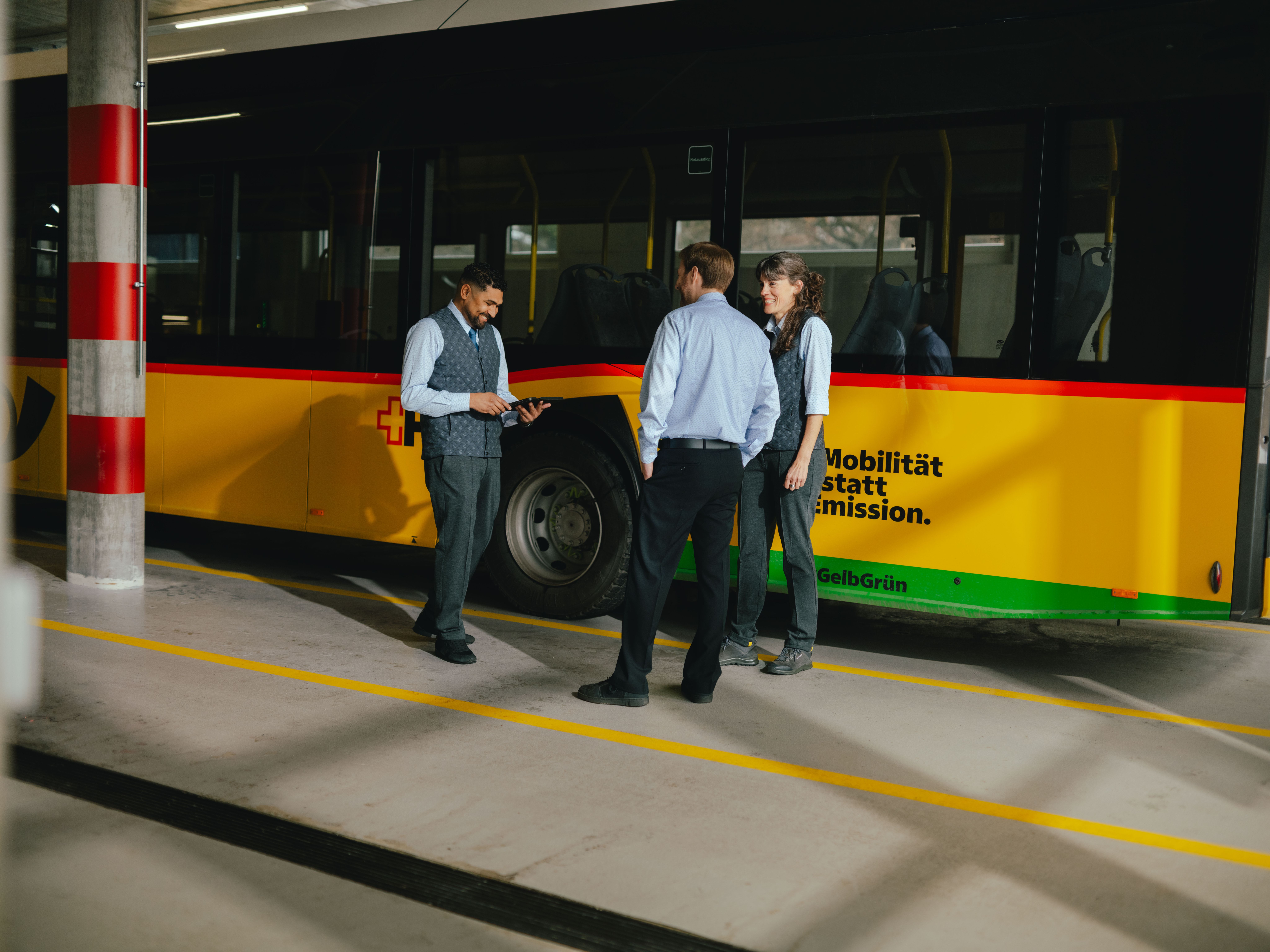 Three people are standing in front of a yellow and black vehicle. One individual is holding a tablet and pointing at the screen, while the other two stand nearby observing. All are wearing formal clothing, such as shirts and vests. The background shows the side of the vehicle with coloured stripes.