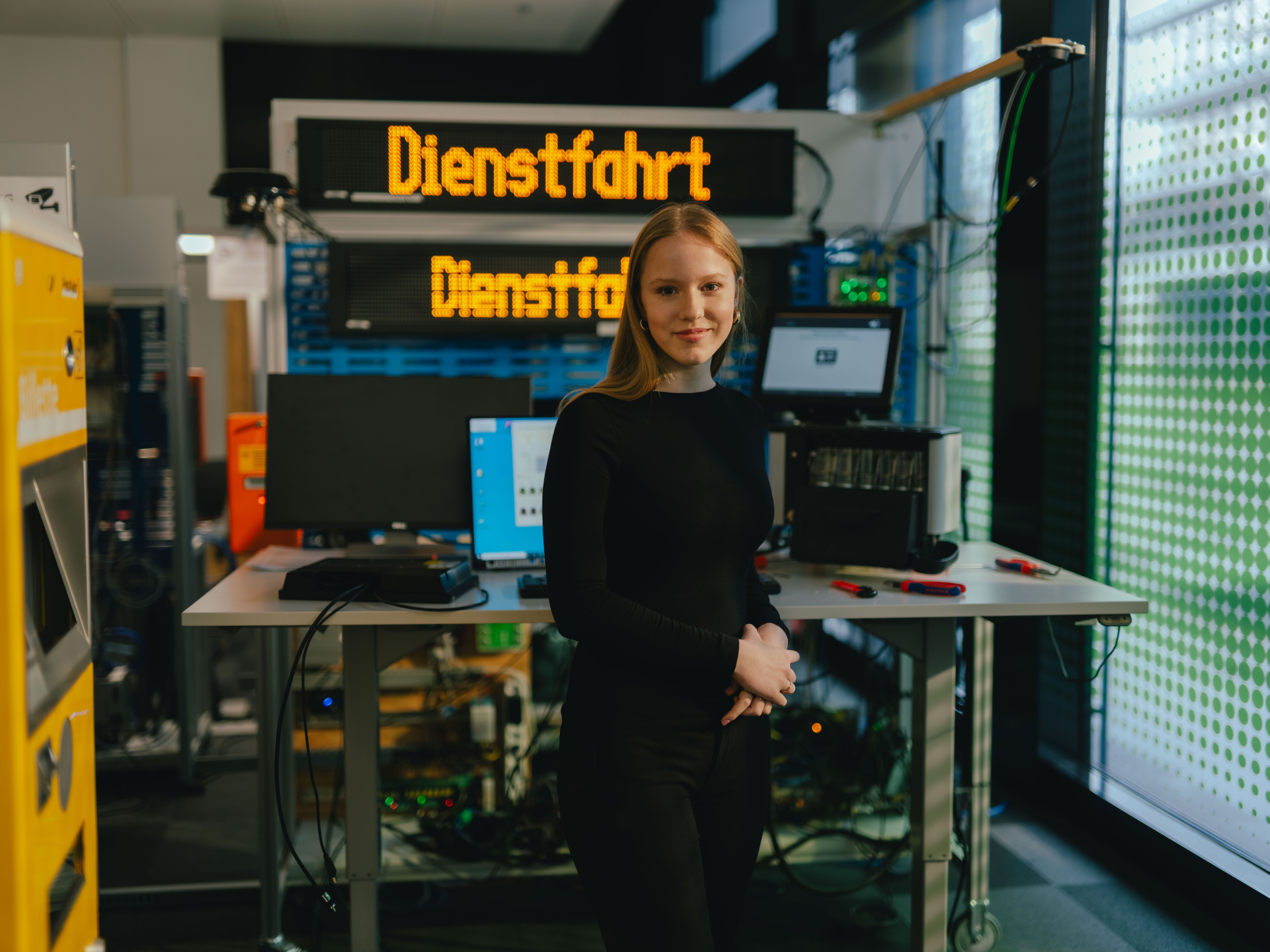 Eine Person steht in einem technischen Arbeitsraum vor einem Tisch mit mehreren Monitoren, Computern und elektronischen Geräten. Hinter der Person befindet sich eine grosse digitale Anzeigetafel mit der gelben Schrift „Dienstfahrt“. Links im Bild sind weitere technische Anlagen und ein gelbes Gerät sichtbar. Der Raum wirkt wie eine Test‑ oder Werkstattumgebung.