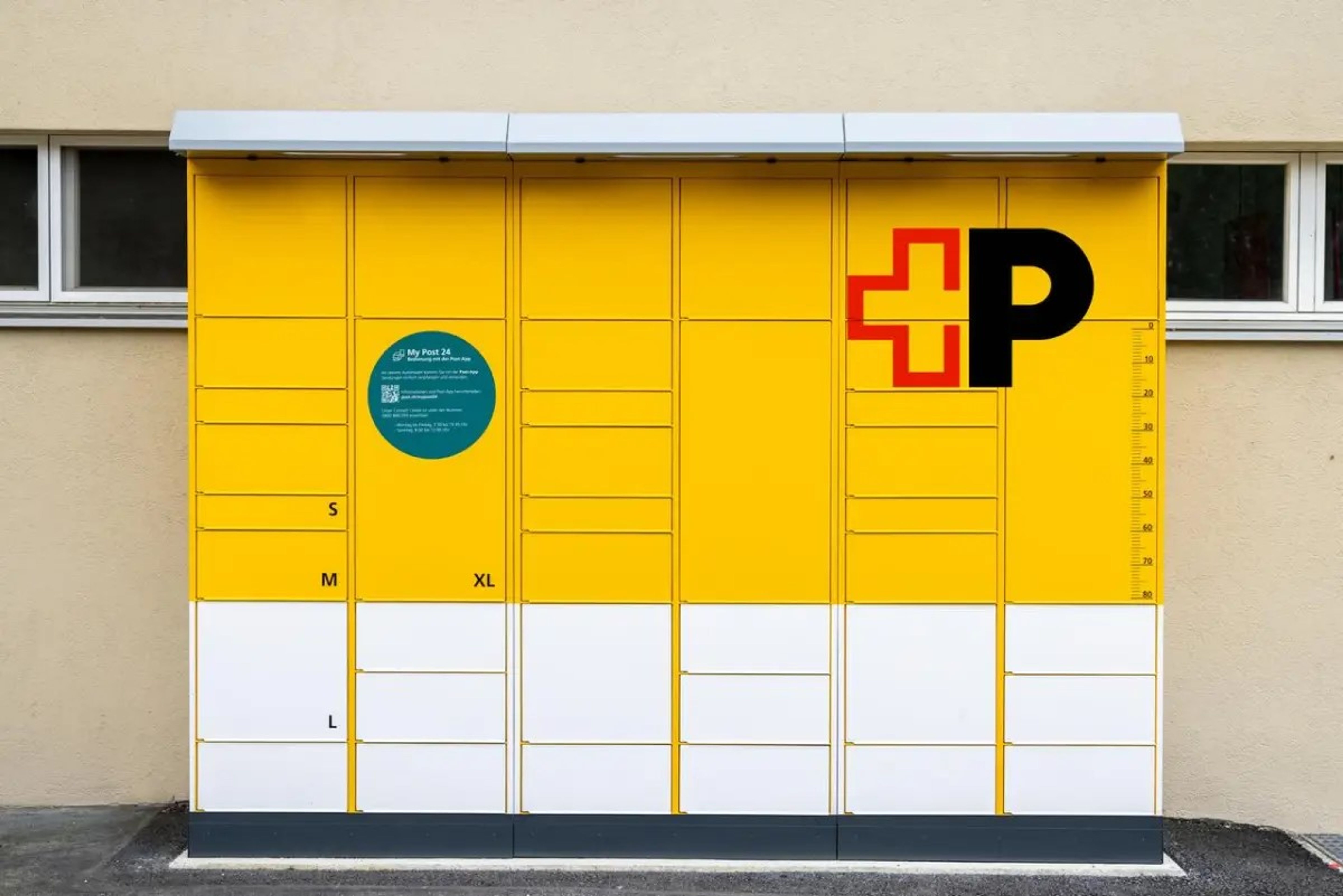 Automate jaune pour colis avec plusieurs compartiments de tailles différentes (S, M, L, XL) et un grand ‘P’ noir avec logo croix rouge. À gauche, un cercle vert avec des instructions d’utilisation. En arrière-plan, un mur beige avec deux fenêtres étroites.