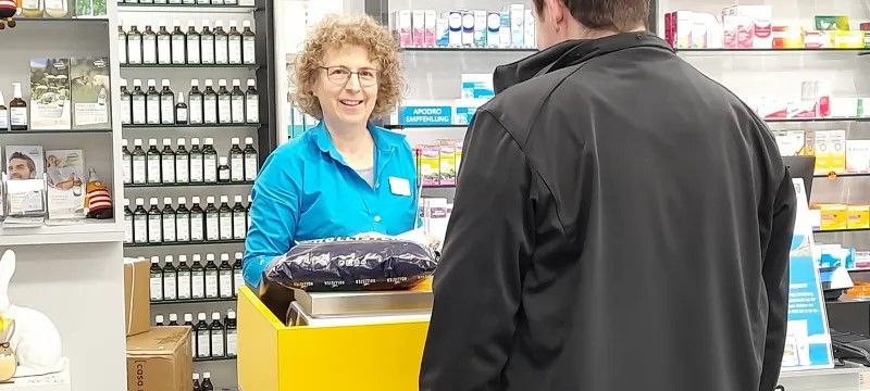 Servizio postale in una farmacia: una persona è al banco postale giallo e consegna un pacco. Sullo sfondo scaffali con medicinali, flaconi e prodotti per la salute.