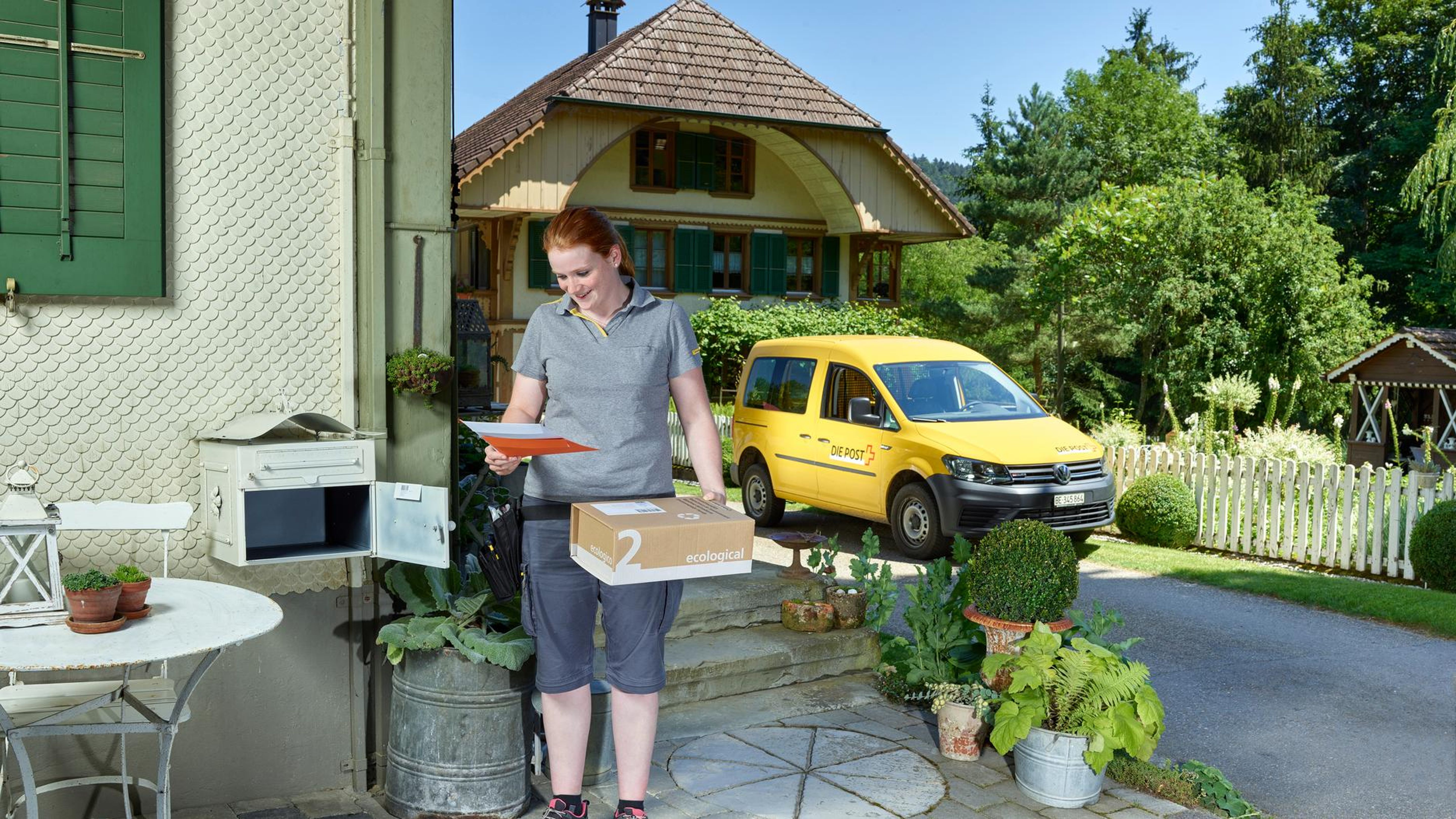 Strassenszene in einem Dorf mit einem alten Gebäude mit Holzverkleidung und roten Fensterläden. Vor dem Haus führt eine Rampe mit Geländer zu einer Tür. Drei große Blumentöpfe stehen am Rand der Straße, im Hintergrund sind weitere Häuser und ein blauer Himmel mit Wolken sichtbar