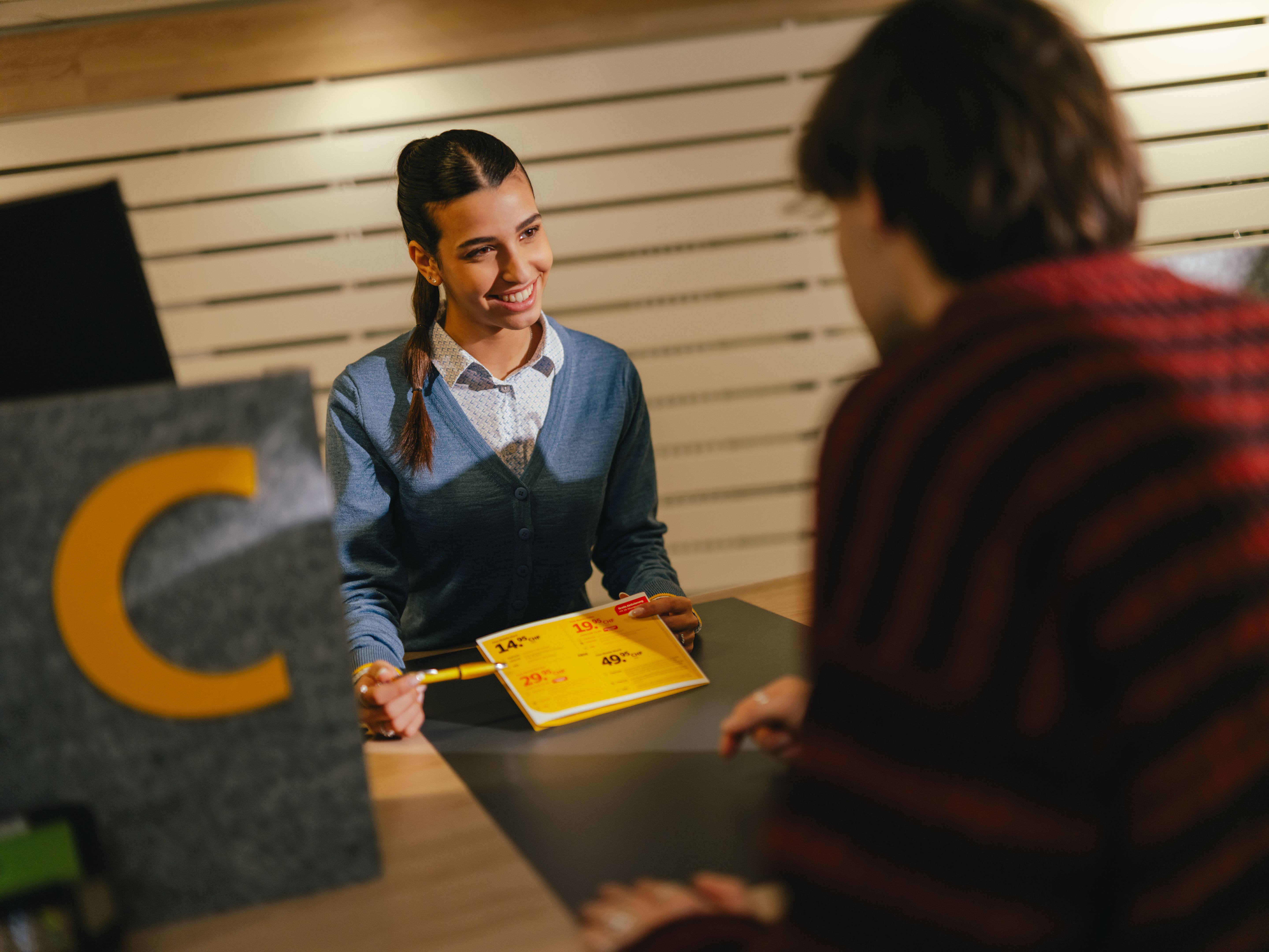 Une personne est assise à un guichet et remet un formulaire jaune à une autre personne au-dessus du comptoir. La zone du comptoir est marquée par un élément gris qui montre une grande lettre jaune «C». L’environnement ressemble à un espace d’accueil ou de service avec des lamelles de bois clair en arrière-plan.