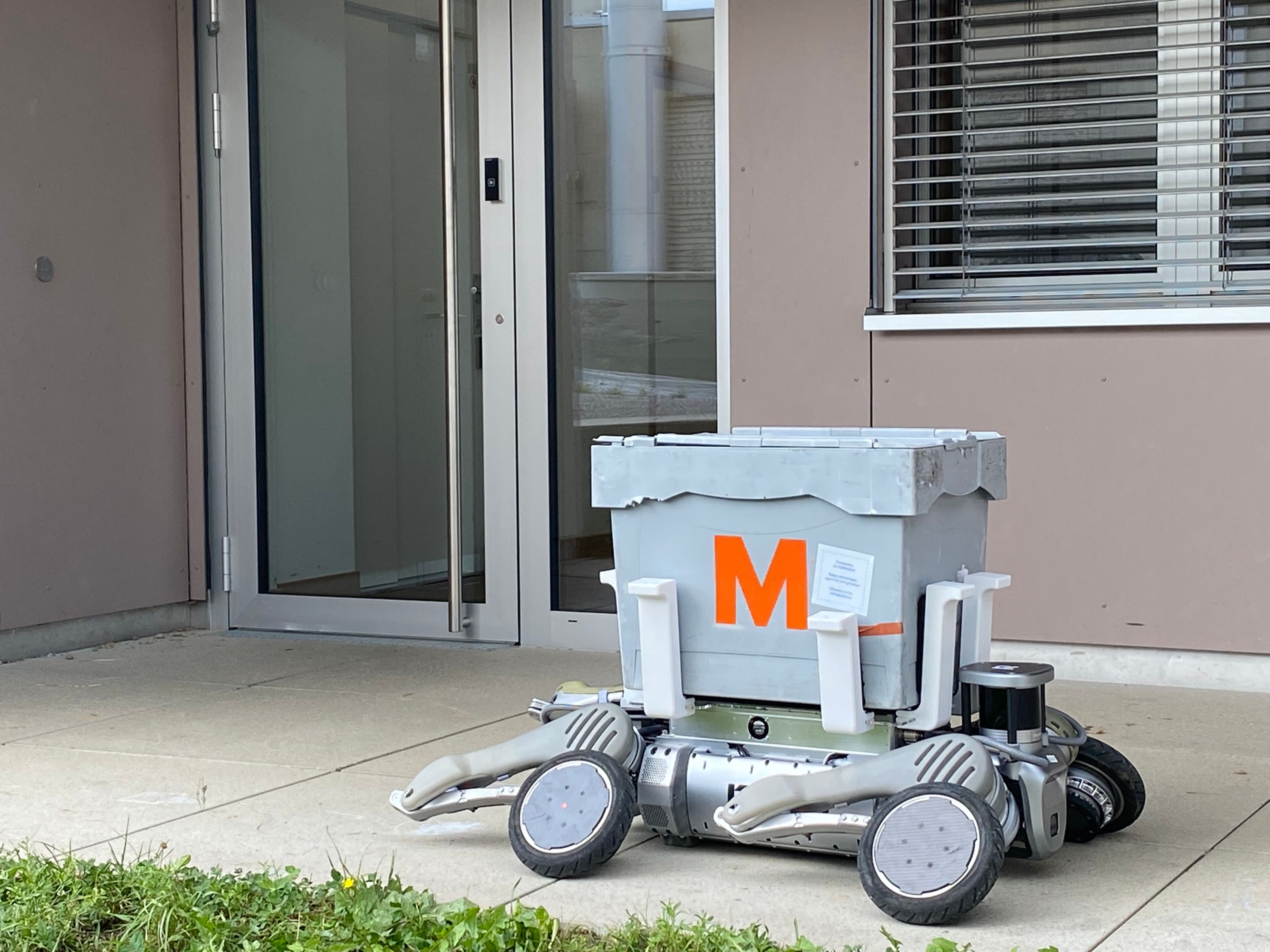 Vierbeiniger Roboter mit grauer Transportbox vor dem Eingang eines Gebäudes mit Glas- und Metalltür sowie Fenster mit Jalousien.