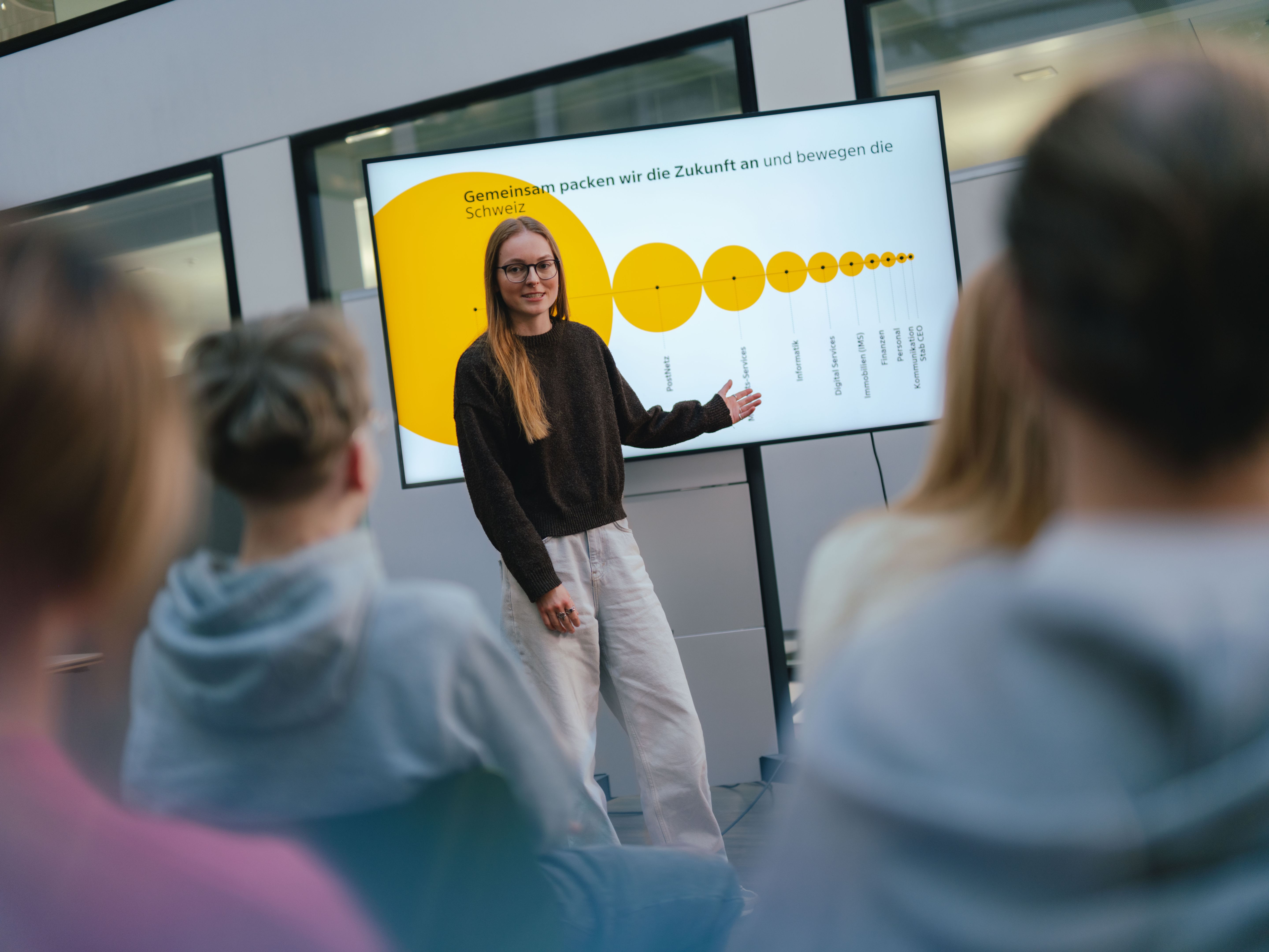 Une personne se tient devant un grand écran et fait une présentation pour un public assis. L’écran affiche un graphique jaune avec des cercles de différentes tailles et des points marqués. La salle est dotée de grandes fenêtres et plusieurs personnes sont visibles de l’arrière-plan.