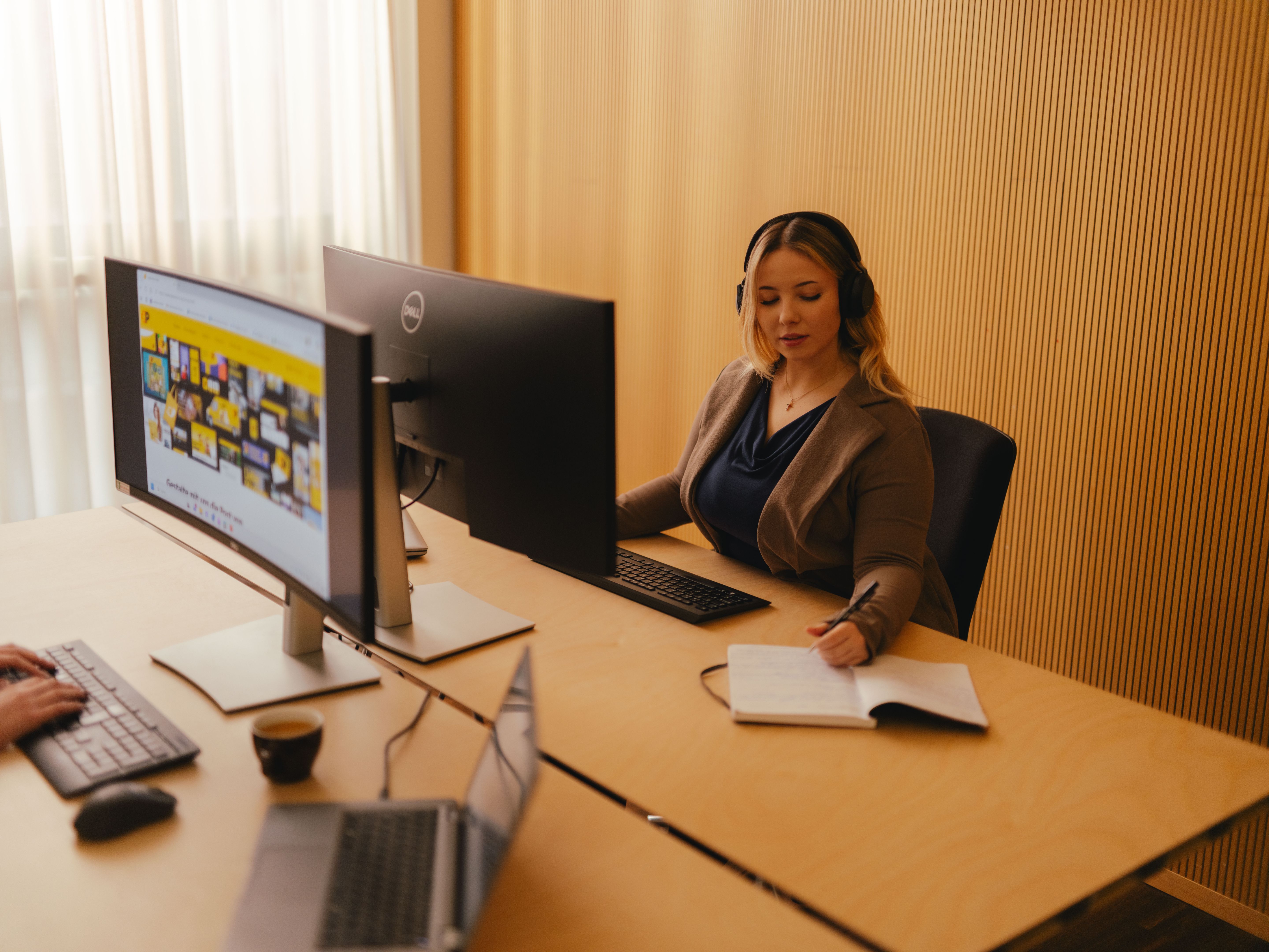 Une personne est assise à un bureau dans un environnement de bureau, utilisant un grand écran et un clavier. Elle porte un casque audio et a un carnet ouvert devant elle. Au premier plan, un autre ordinateur portable est visible sur une seconde table. La pièce comporte de grands rideaux clairs et un mur au motif vertical.