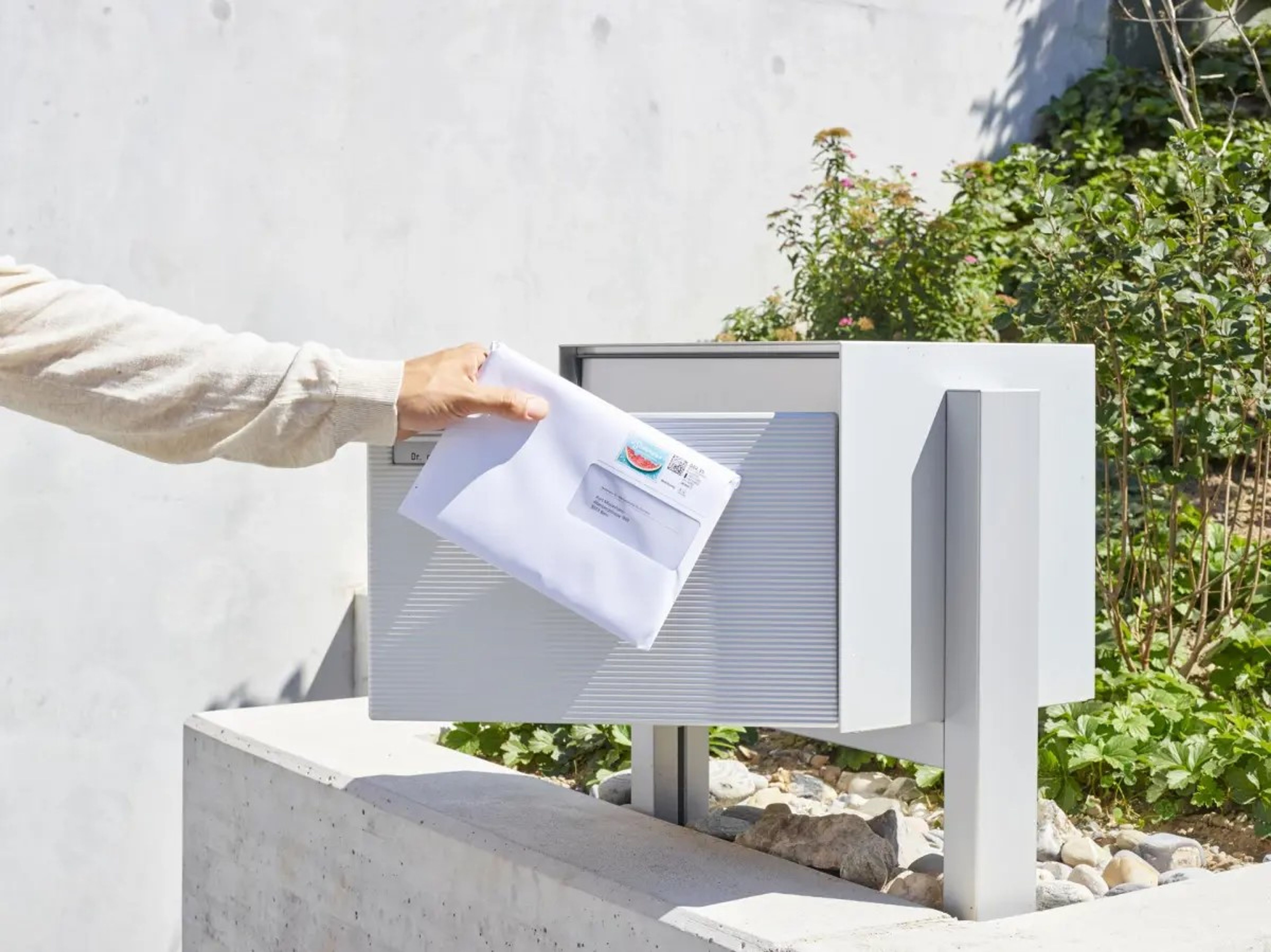Hand steckt einen weissen Brief in einen modernen, grauen Briefkasten im Freien. Im Hintergrund sind Pflanzen und eine Betonmauer sichtbar.