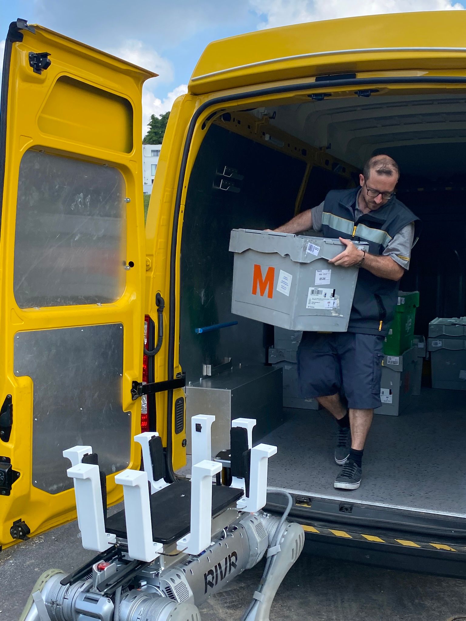 Geöffneter gelber Post-Transporter, eine Person entlädt eine graue Transportbox mit rotem M. Vor dem Fahrzeug steht ein vierbeiniger Roboter bereit.
