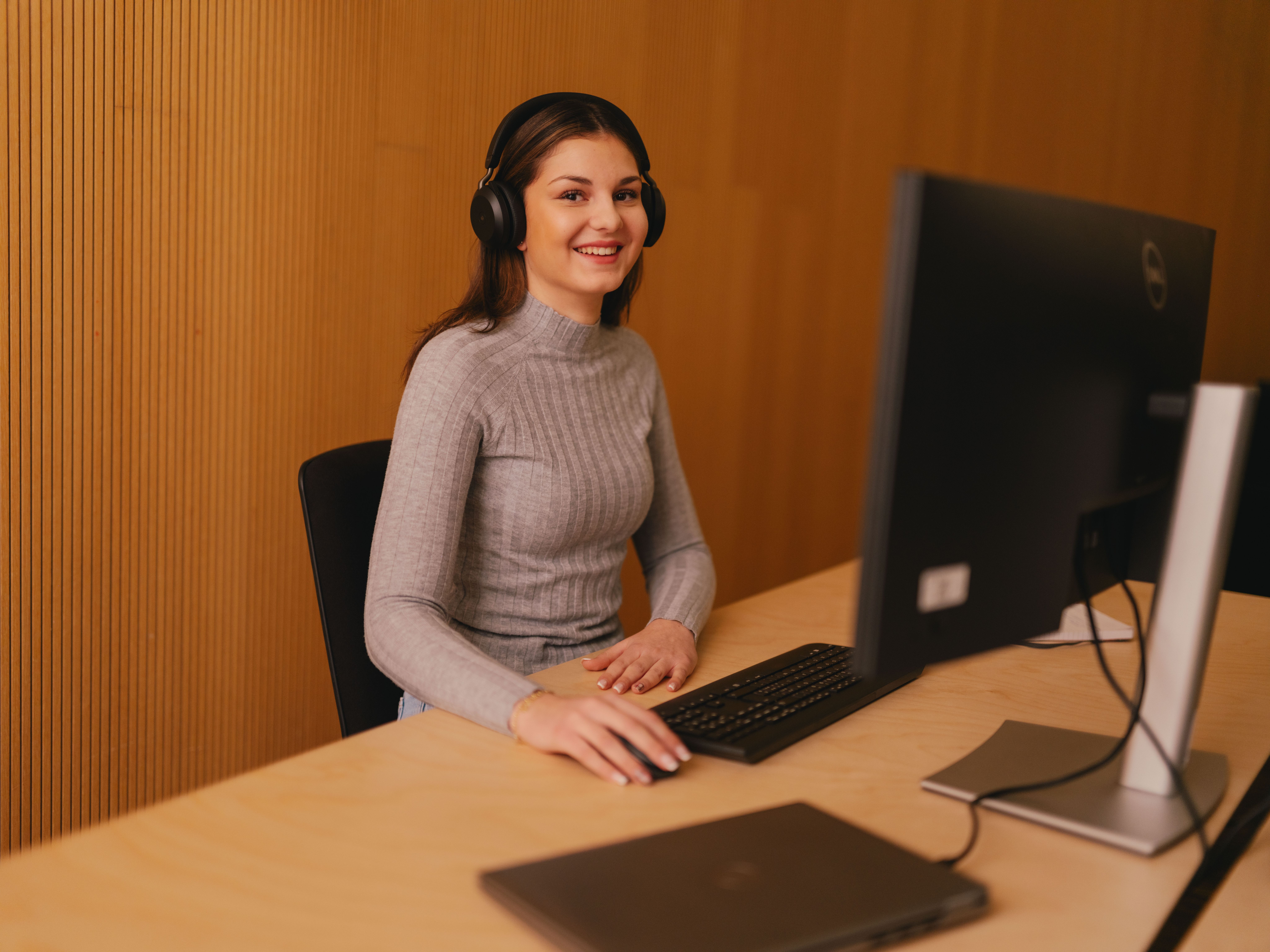 Una persona siede a una scrivania e lavora a un computer desktop. La persona sta usando grandi cuffie over-ear e indossa una maglia a righe a maniche lunghe. Davanti a questa persona ci sono una tastiera e un mouse. Sulla scrivania c’è anche un computer portatile chiuso. Lo sfondo è costituito da pannelli in legno verticali.