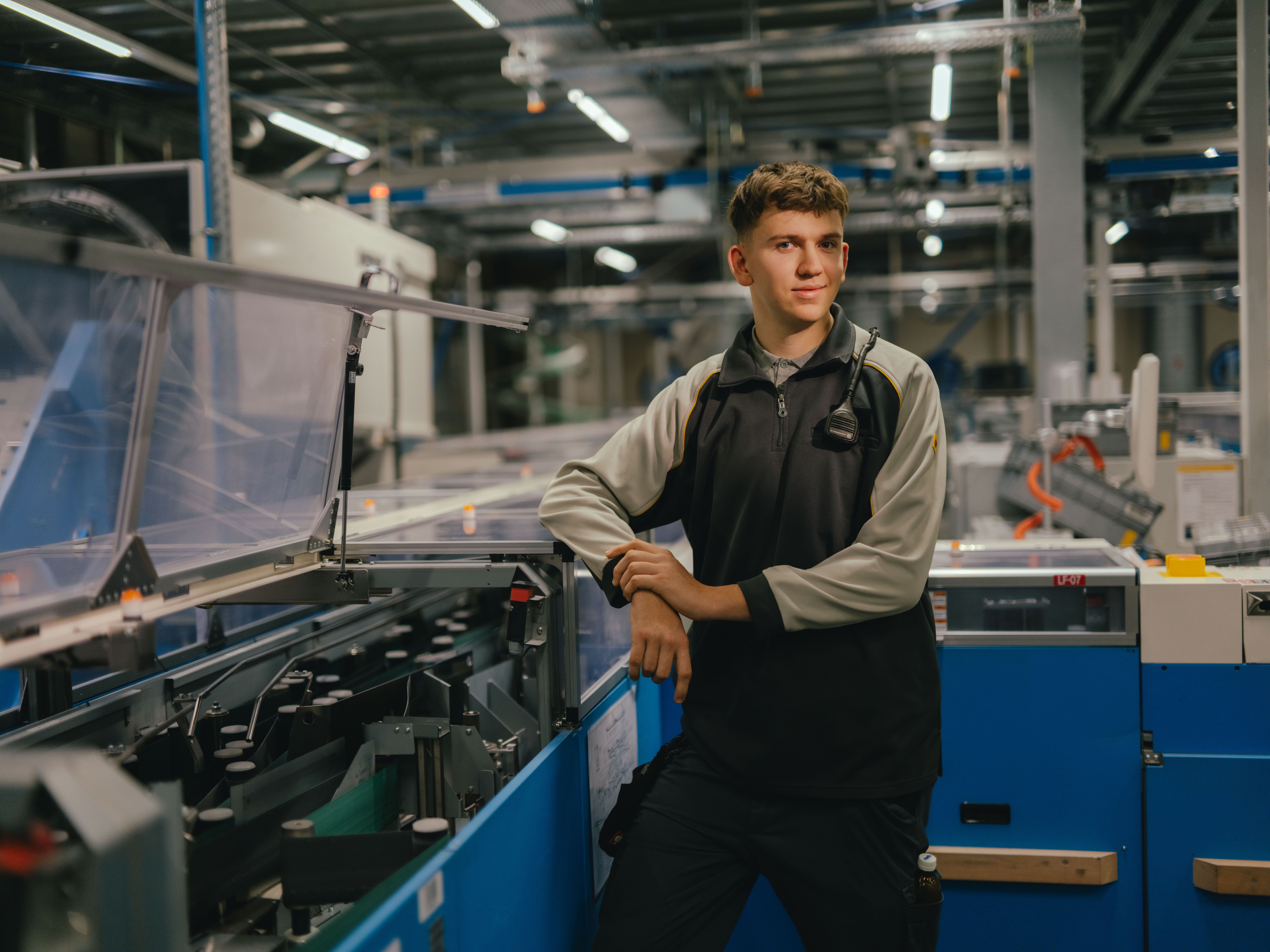 Une personne se tient dans une halle industrielle et s’appuie avec ses deux bras sur une machine équipée de capots transparents. La personne porte une veste de travail bicolore. En arrière-plan, d’autres machines, structures métalliques et éléments d’éclairage de l’environnement de production sont visibles.