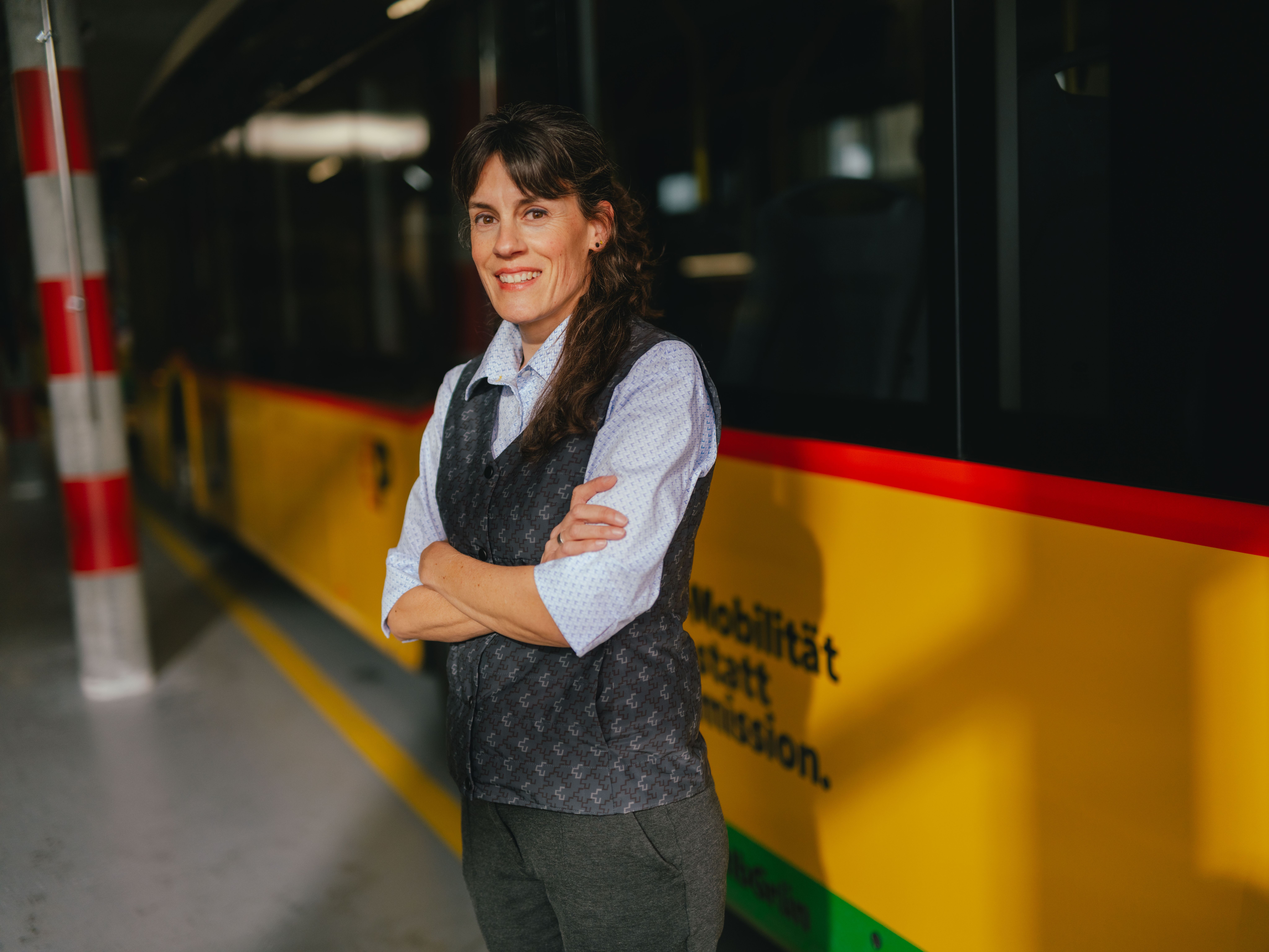 Une personne se tient les bras croisés dans un hall, à côté d’un véhicule jaune et noir. La personne porte une chemise claire et un gilet foncé. L’environnement ressemble à un dépôt ou une zone de stationnement, avec des colonnes rayées et un sol réfléchissant.