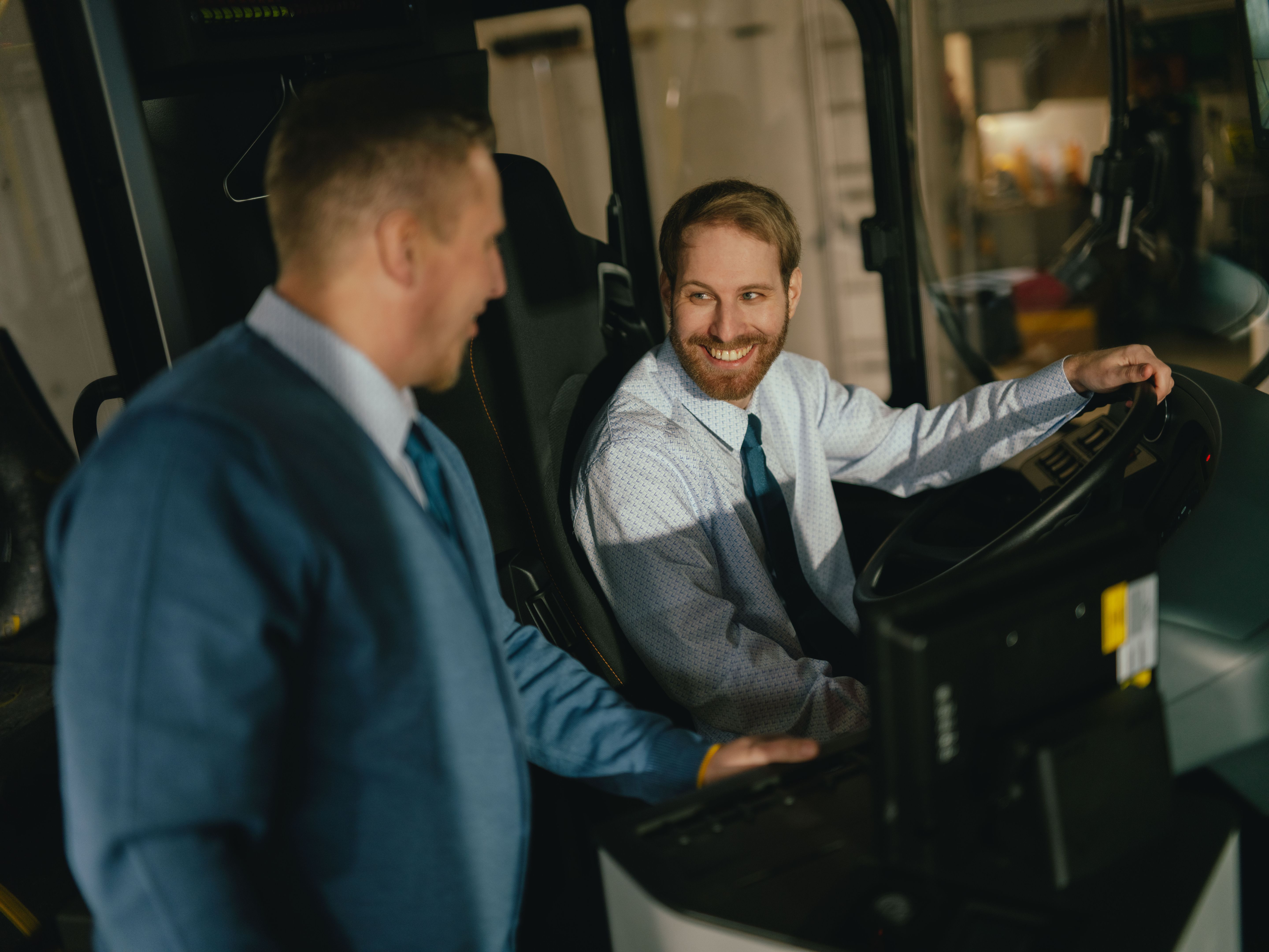 Two people are positioned in the driver’s area of a vehicle. One person is seated at the wheel, while the other stands beside them and leans slightly forward. Both are wearing formal clothing. The steering wheel, control elements, and part of the dashboard are visible. The setting appears to be the interior of a bus.