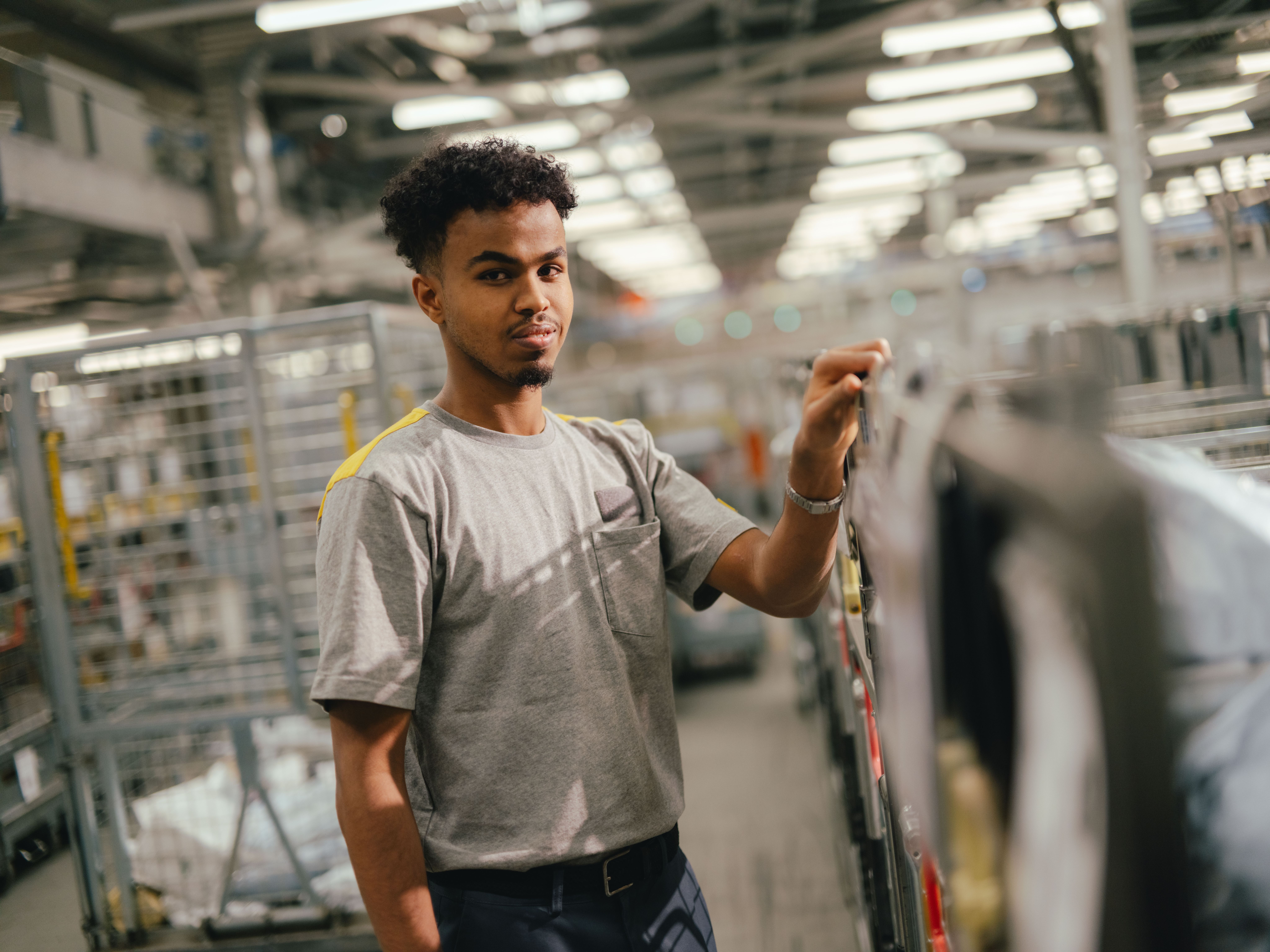 A person is standing in a large hall between metal shelves and transport trolleys. They are wearing a light T‑shirt and holding a handle on a trolley or frame with one hand. The hall is brightly lit, and numerous industrial structures are visible in the background.