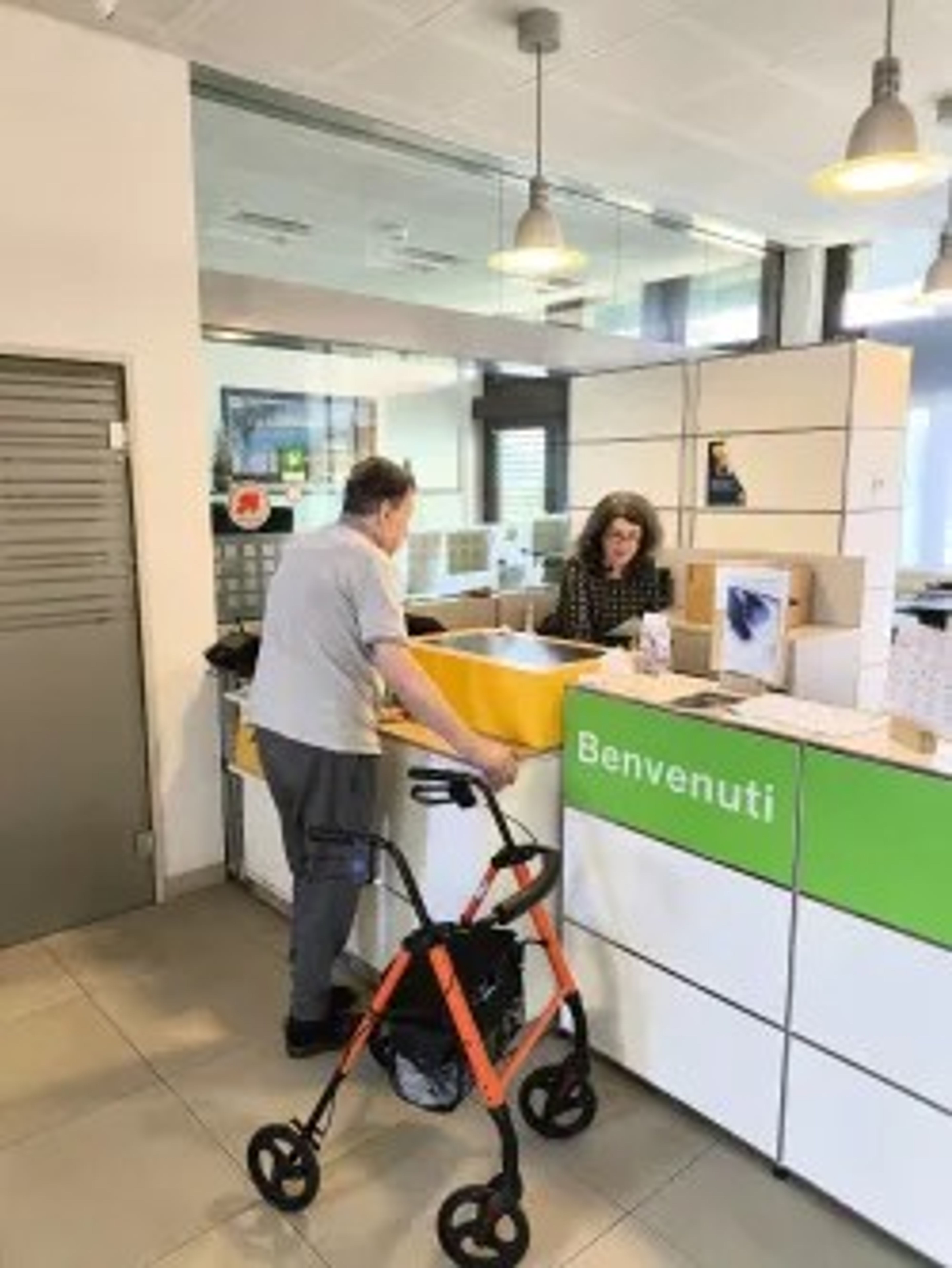 Una persona è davanti a un banco di accoglienza in un edificio moderno. Davanti a sé c’è un deambulatore arancione. Dietro il banco, un’altra persona lavora al computer. Sul banco si vede un grande contenitore giallo. Sulla parte frontale del banco è scritto “Benvenuti”.