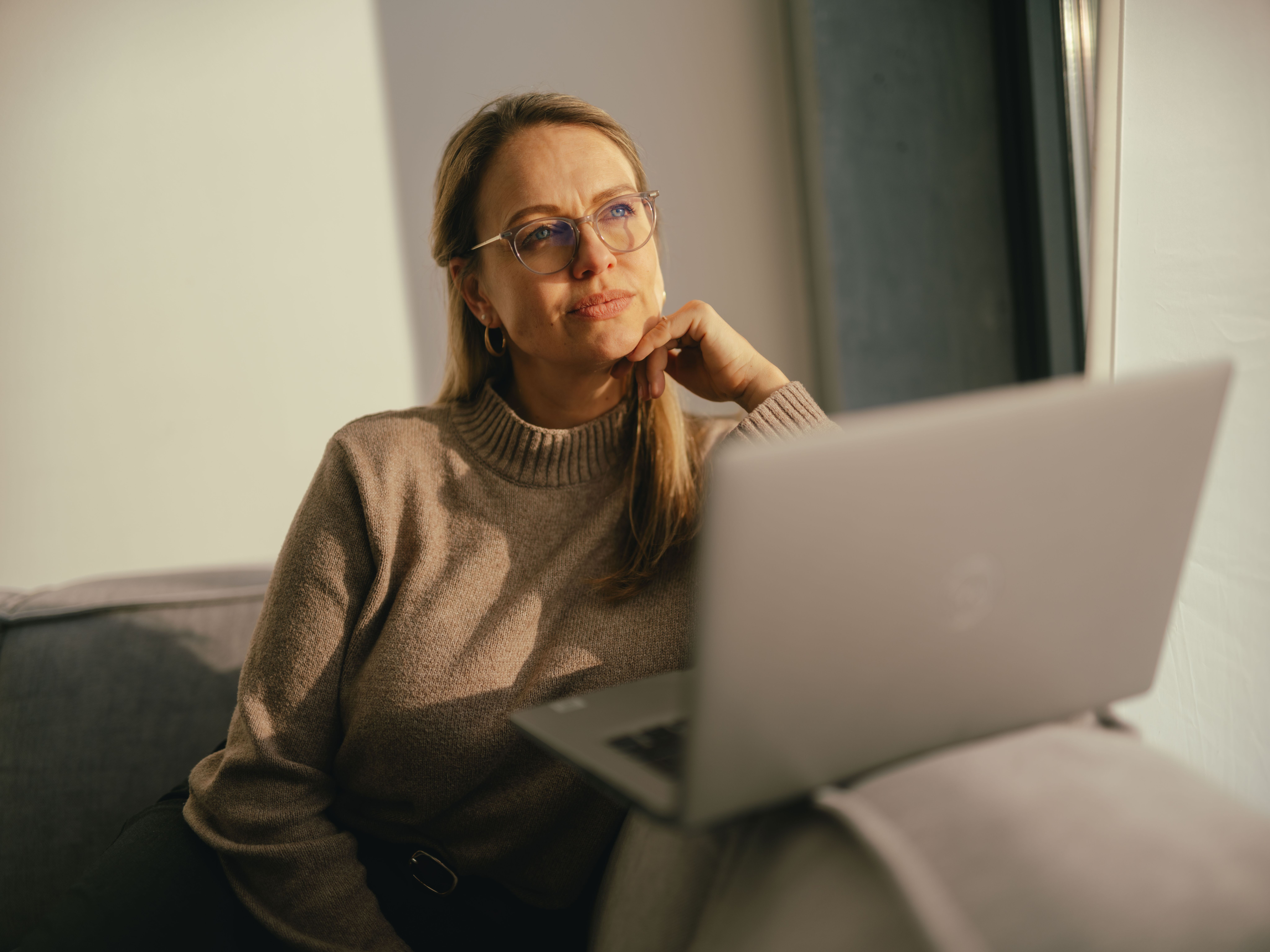 Una persona è seduta su un divano vicino a una finestra e lavora su un computer portatile aperto. La persona indossa un maglione beige, e una luce solare calda illumina l’ambiente.