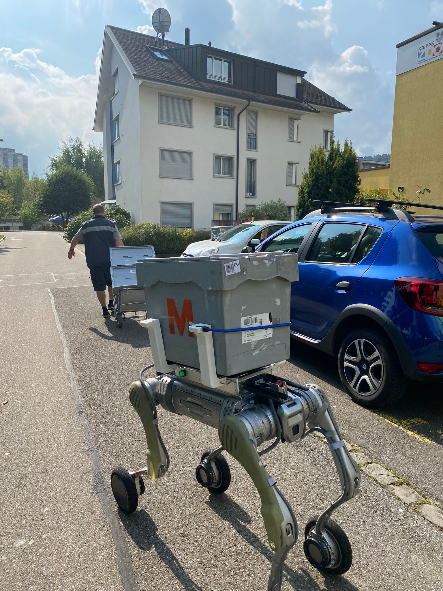 Vierbeiniger Roboter mit Post-Transportbox auf einer Straße, neben geparkten Autos. Im Hintergrund ein mehrstöckiges Wohnhaus und eine Person, die eine weitere Box bewegt.