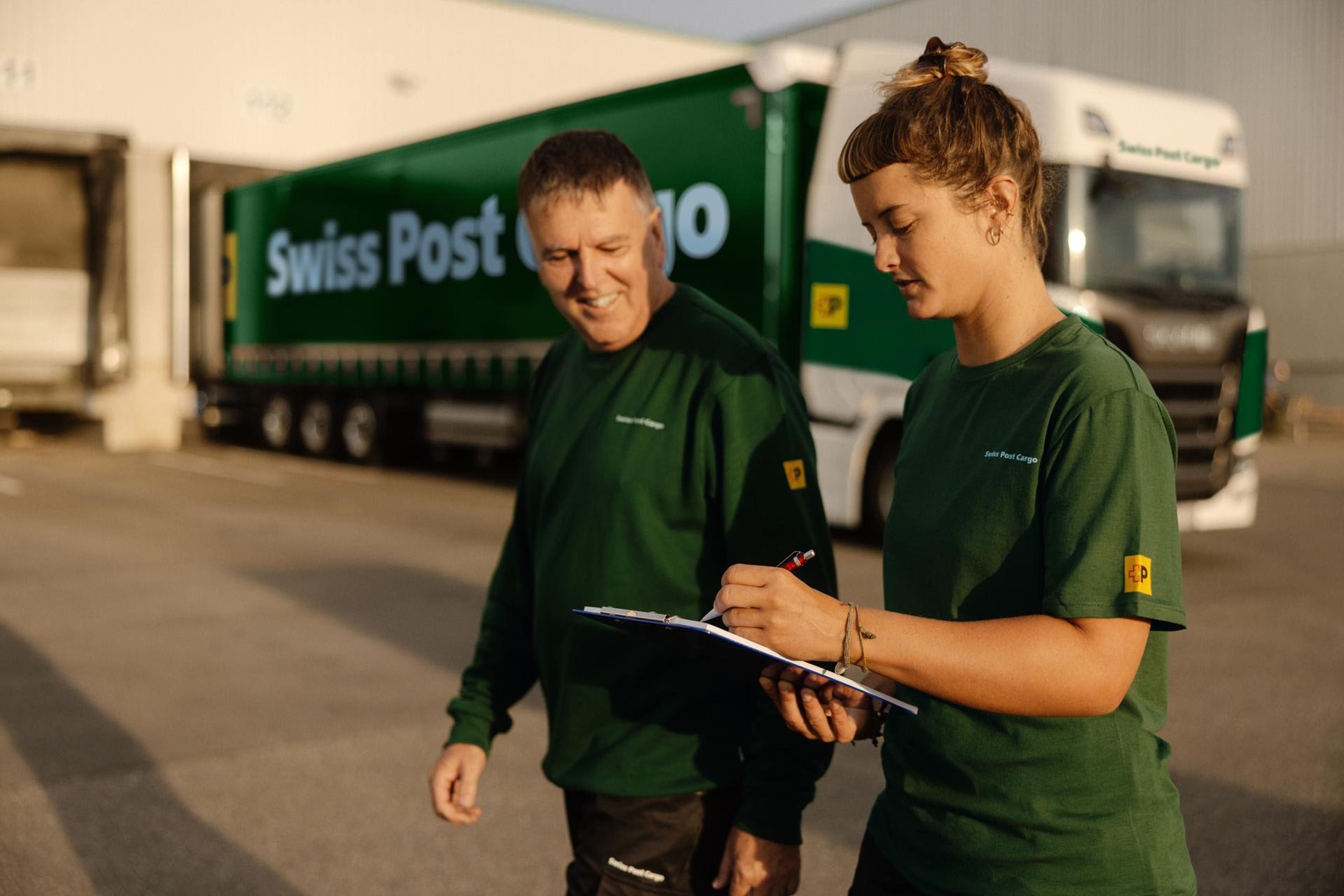 Deux personnes employées de Swiss Post Cargo passent devant un camion Swiss Post Cargo stationné à un quai de chargement.
