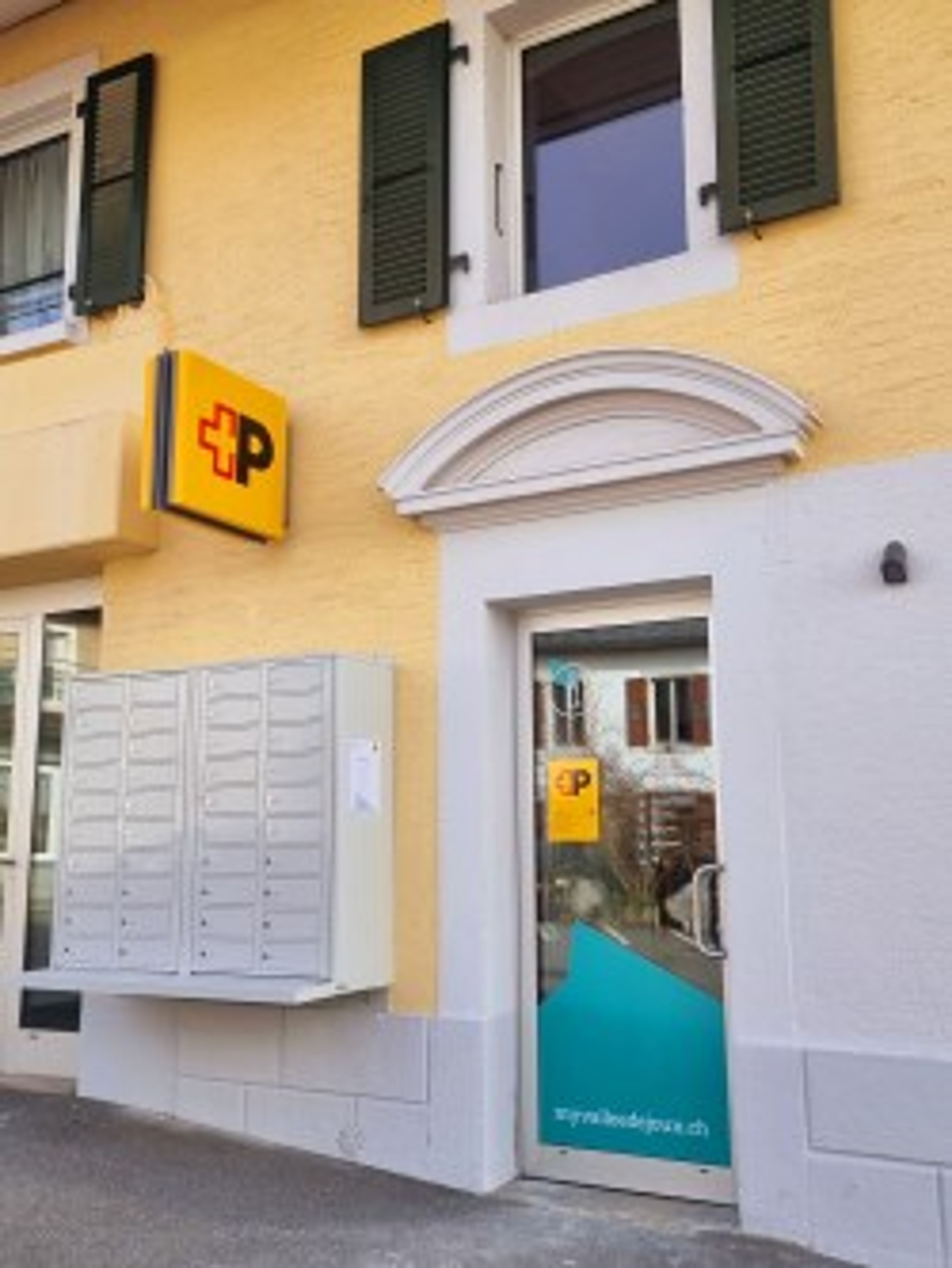 Ingresso di un ufficio postale in un edificio giallo con persiane grigie. A sinistra una fila di caselle postali, a destra una porta a vetri con il logo della Posta e l’URL “mypostservice.ch”. Sopra la porta un arco decorativo bianco.