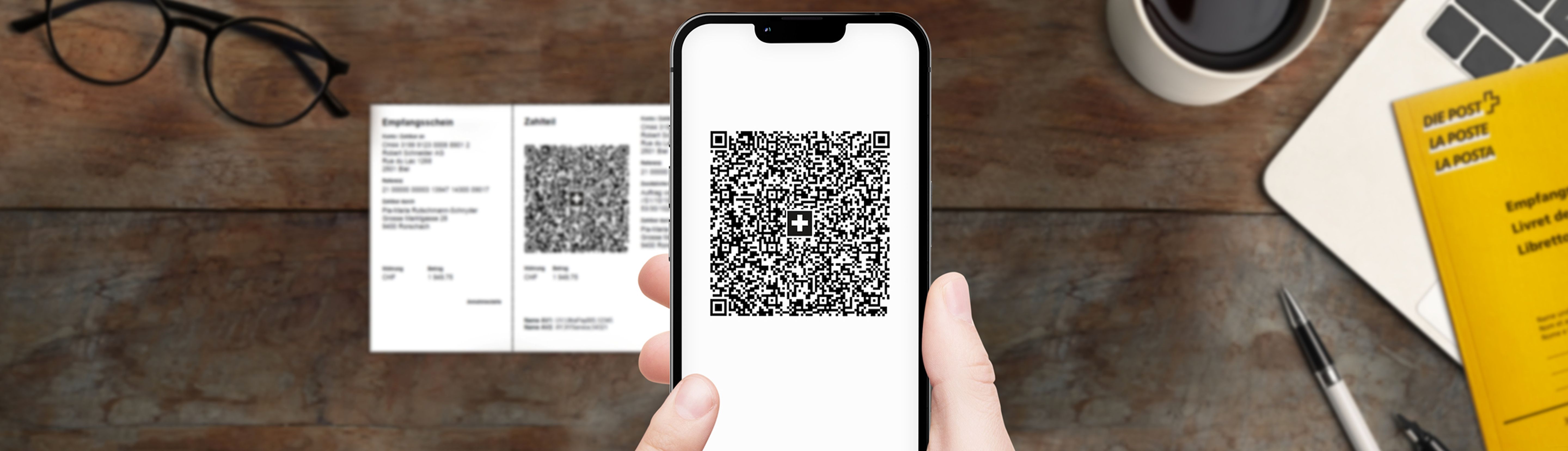 Hand hält ein Smartphone mit einem QR-Code auf dem Bildschirm. Auf dem Tisch liegen eine QR-Rechnung, eine Brille, ein gelbes Postheft, ein Laptop, ein Stift und eine Kaffeetasse.