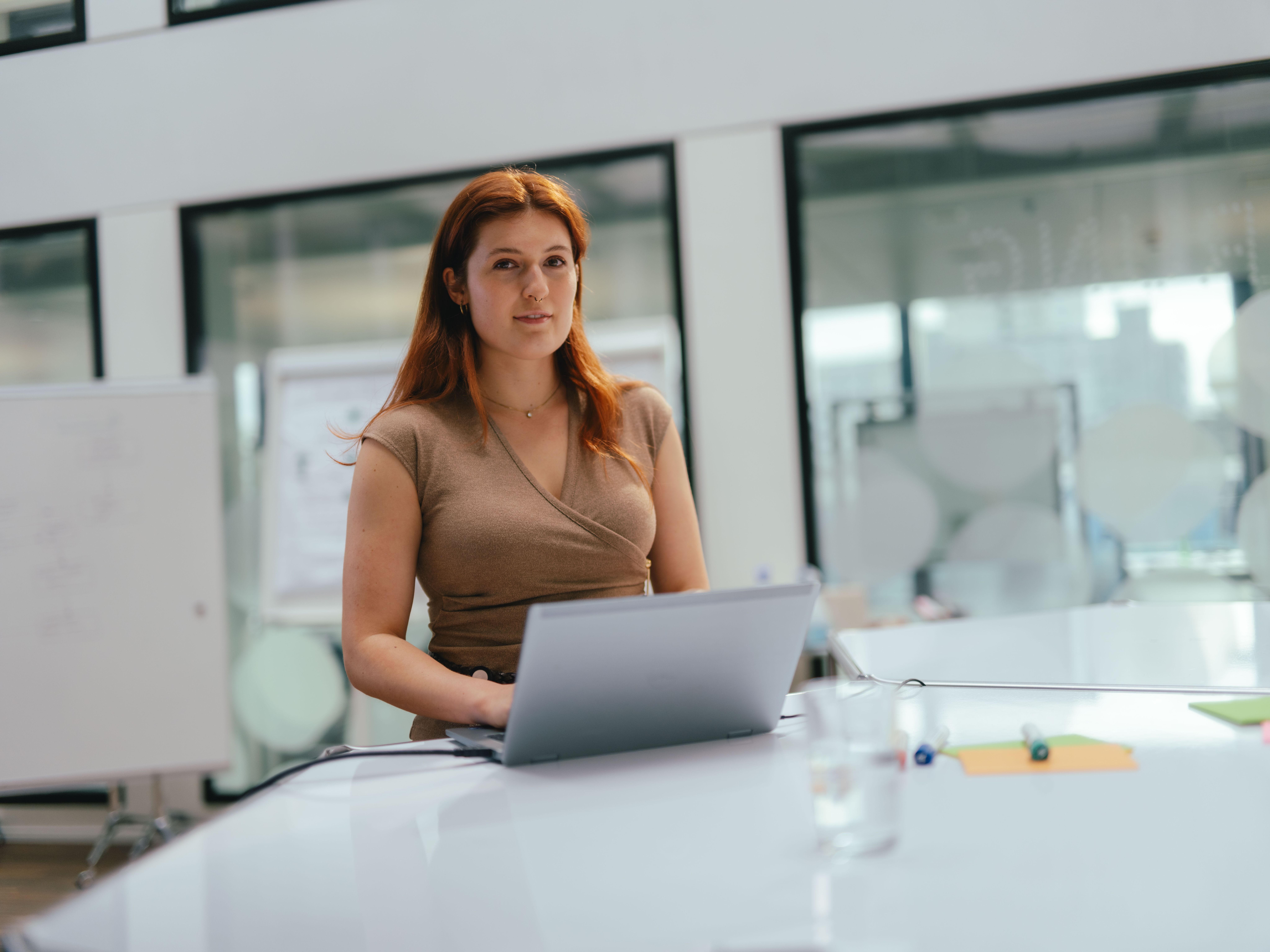 Eine Person sitzt an einem grossen, weissen Konferenztisch in einem modernen Büro. Vor der Person steht ein geöffnetes Laptop. Im Hintergrund sind Glaswände, ein Whiteboard und weitere Büroelemente sichtbar. Auf dem Tisch liegen ein paar Stifte und eine Wasserflasche.