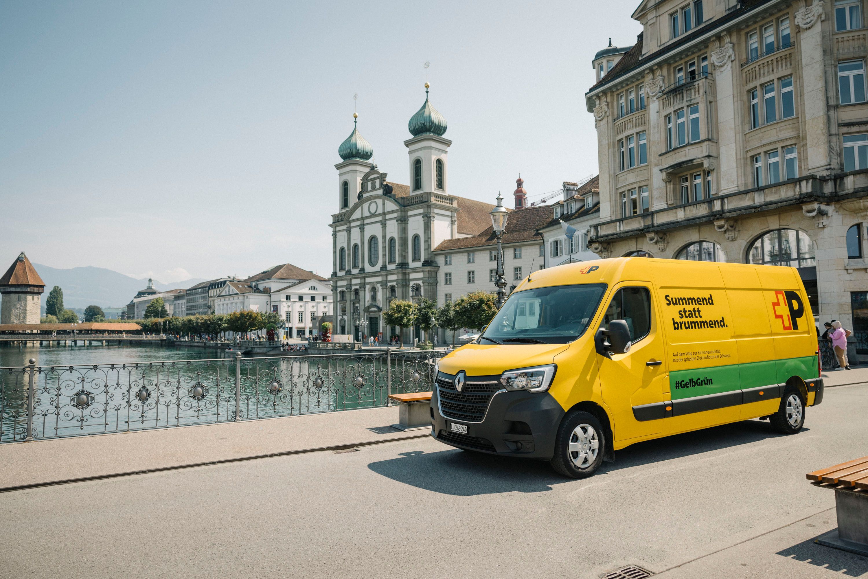 A yellow Swiss Post delivery van is parked on a riverside promenade in Lucerne. In the background are the Jesuit Church, historic buildings, and the Reuss River. Text on the van reads: “Summend statt brummend.”.