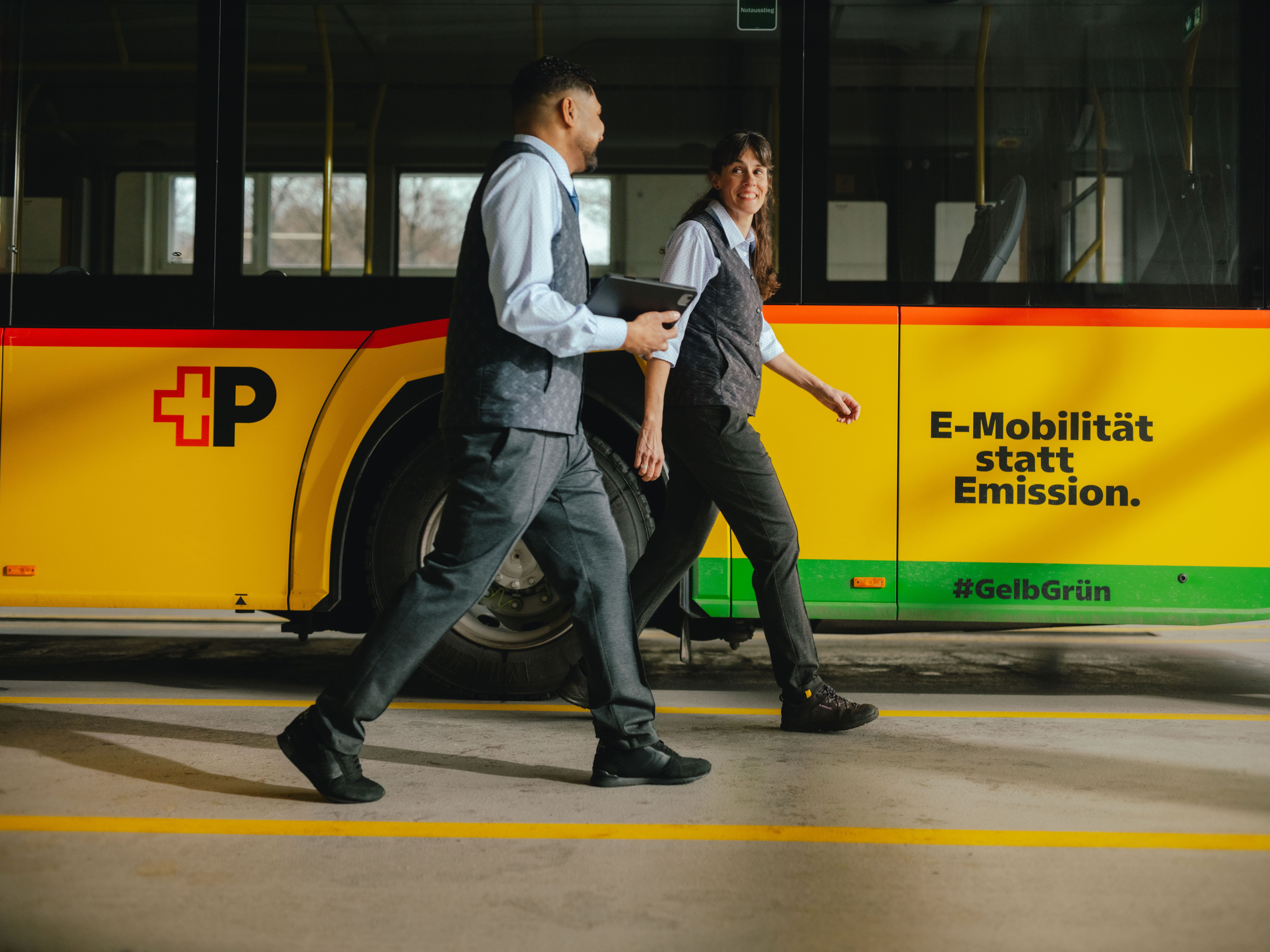 Zwei Personen gehen nebeneinander in einer Halle an einem gelb‑schwarzen Bus vorbei. Eine der Personen hält ein Tablet in der Hand. Auf der Busseite sind der Schriftzug „E‑Mobilität statt Emission.“ sowie Hashtags und farbige Streifen sichtbar. Die Umgebung wirkt wie ein Depot oder eine Servicehalle.