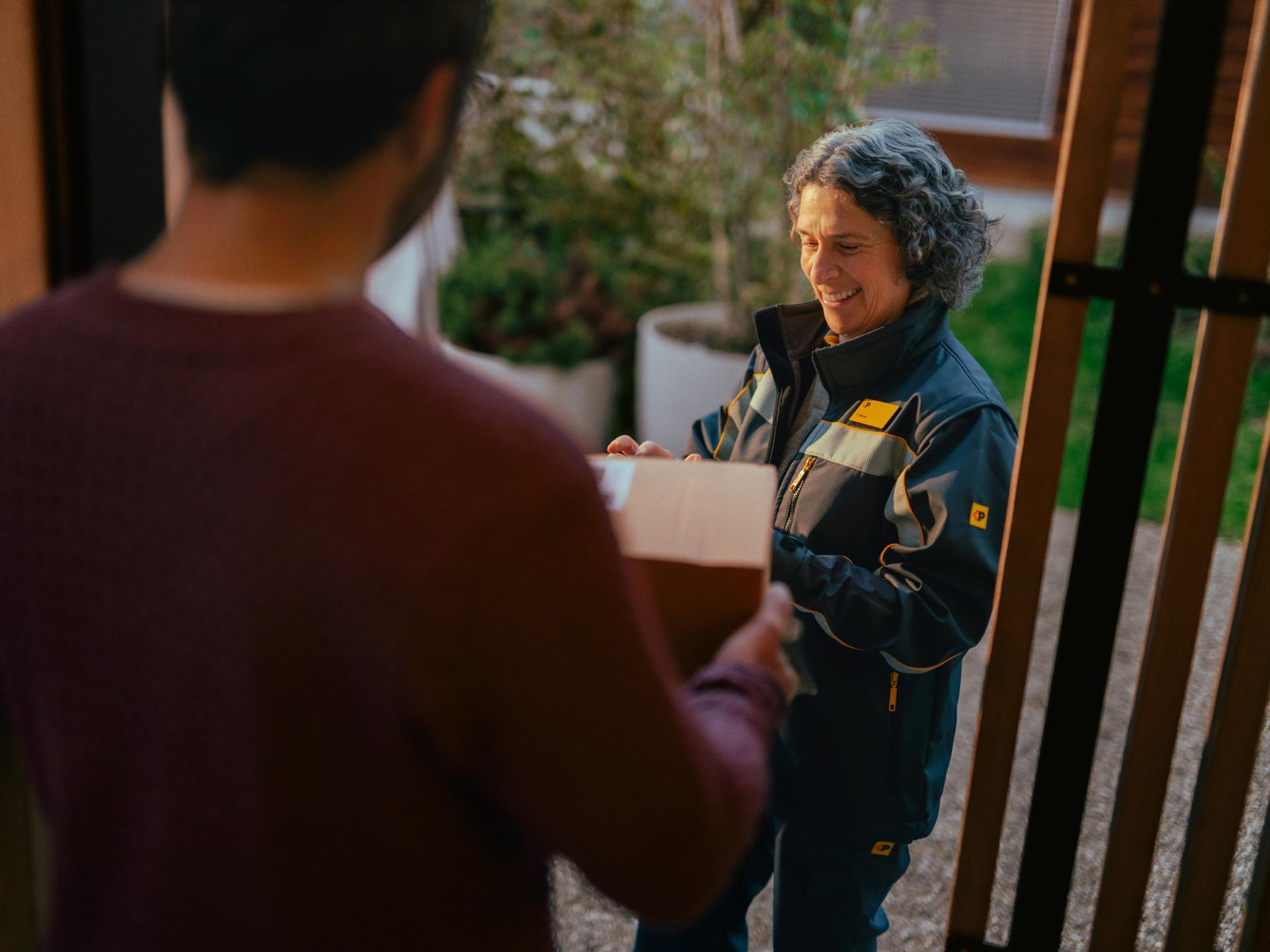 Eine Person in einer Zustelluniform überreicht ein Paket an eine andere Person, die im Vordergrund steht. Die Übergabe findet an einer Haustür statt, mit Pflanzen und einem Holzelement im Hintergrund.