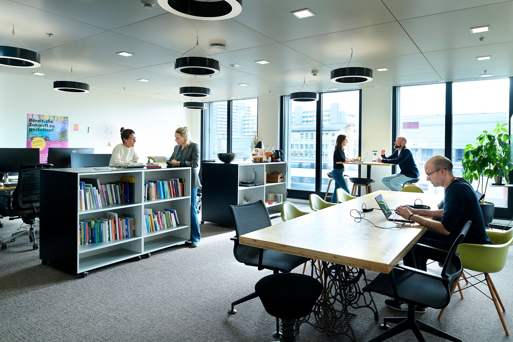 Several people work in a modern, bright office with large windows and books.
