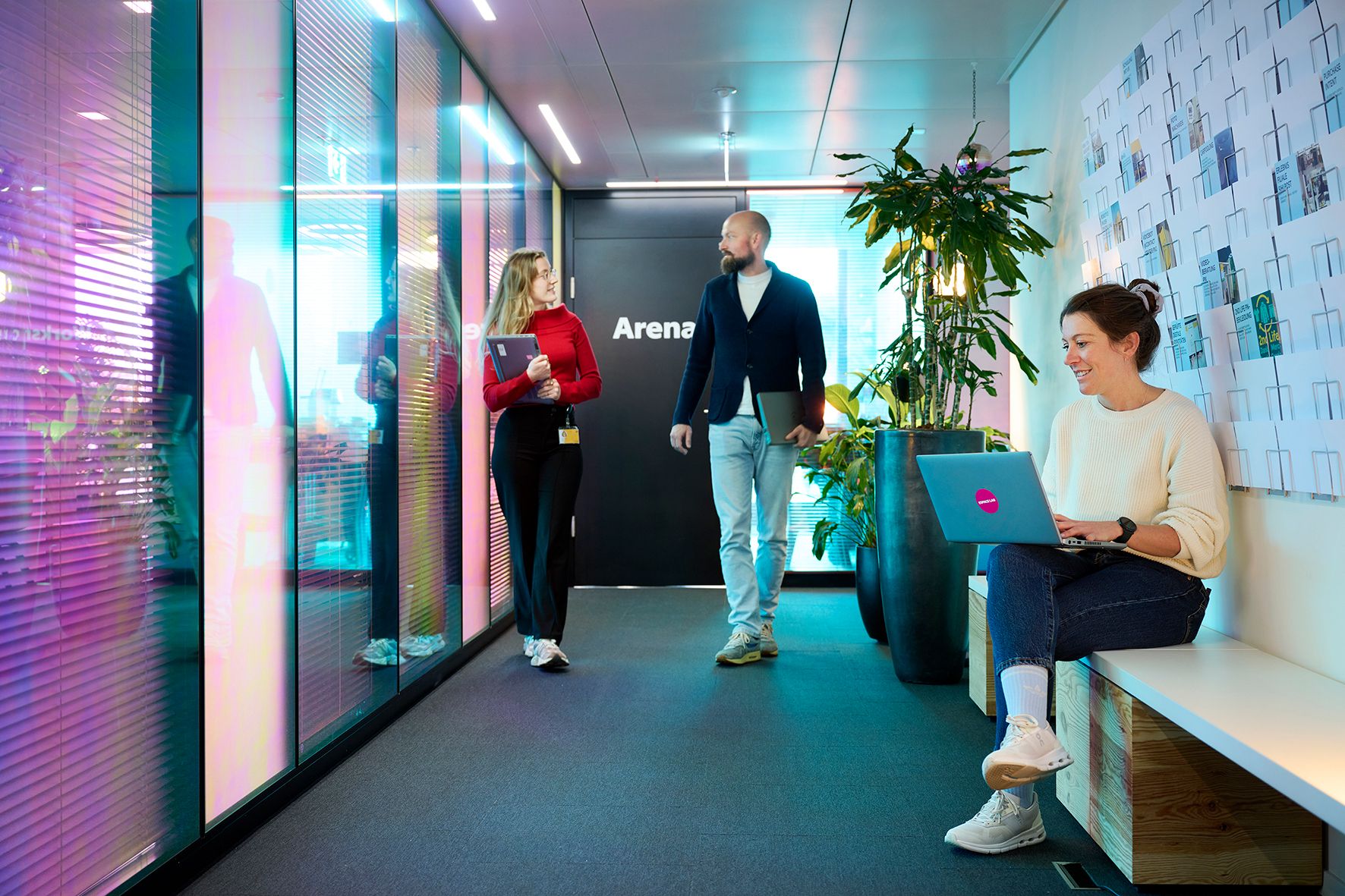 People in a modern office building, walking two, one sitting with a laptop.