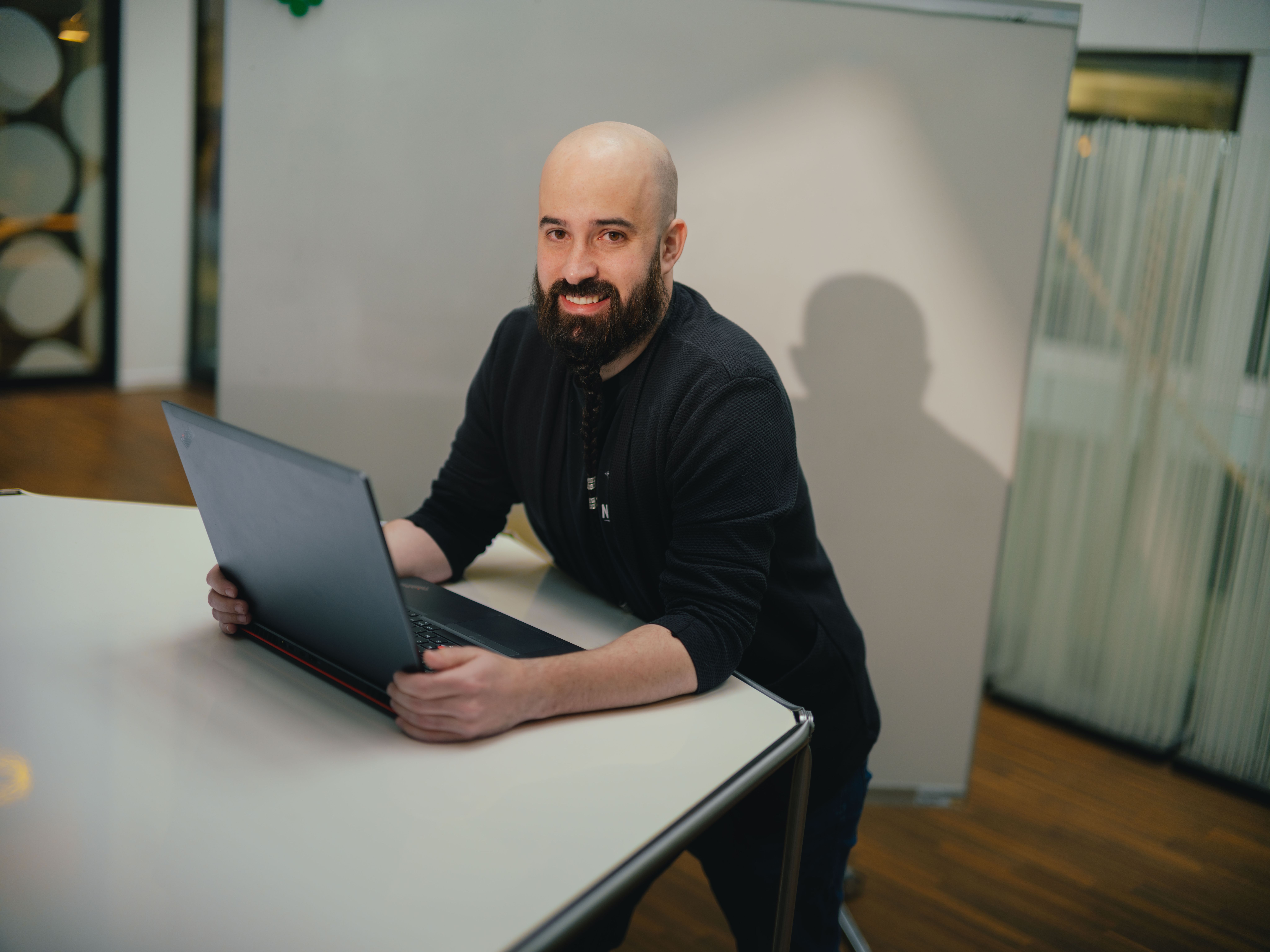 Une personne est assise à une table et travaille sur un ordinateur portable ouvert. La personne porte des vêtements foncés. À l’arrière‑plan, on voit une paroi vitrée avec des structures verticales ainsi qu’une autre zone du bâtiment. La table est claire et présente une surface lisse.