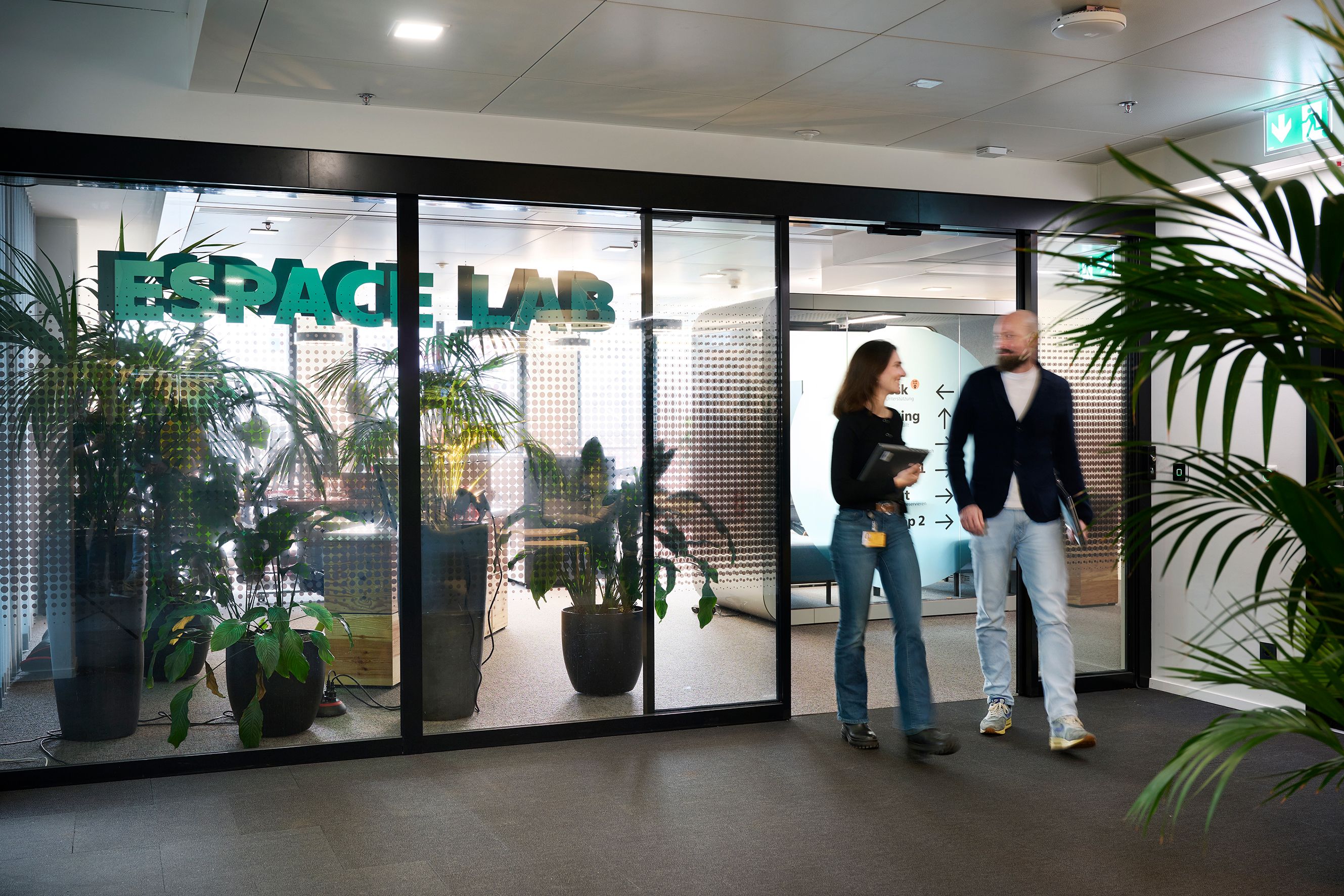 Two people walk past a glass door with plants in the background.