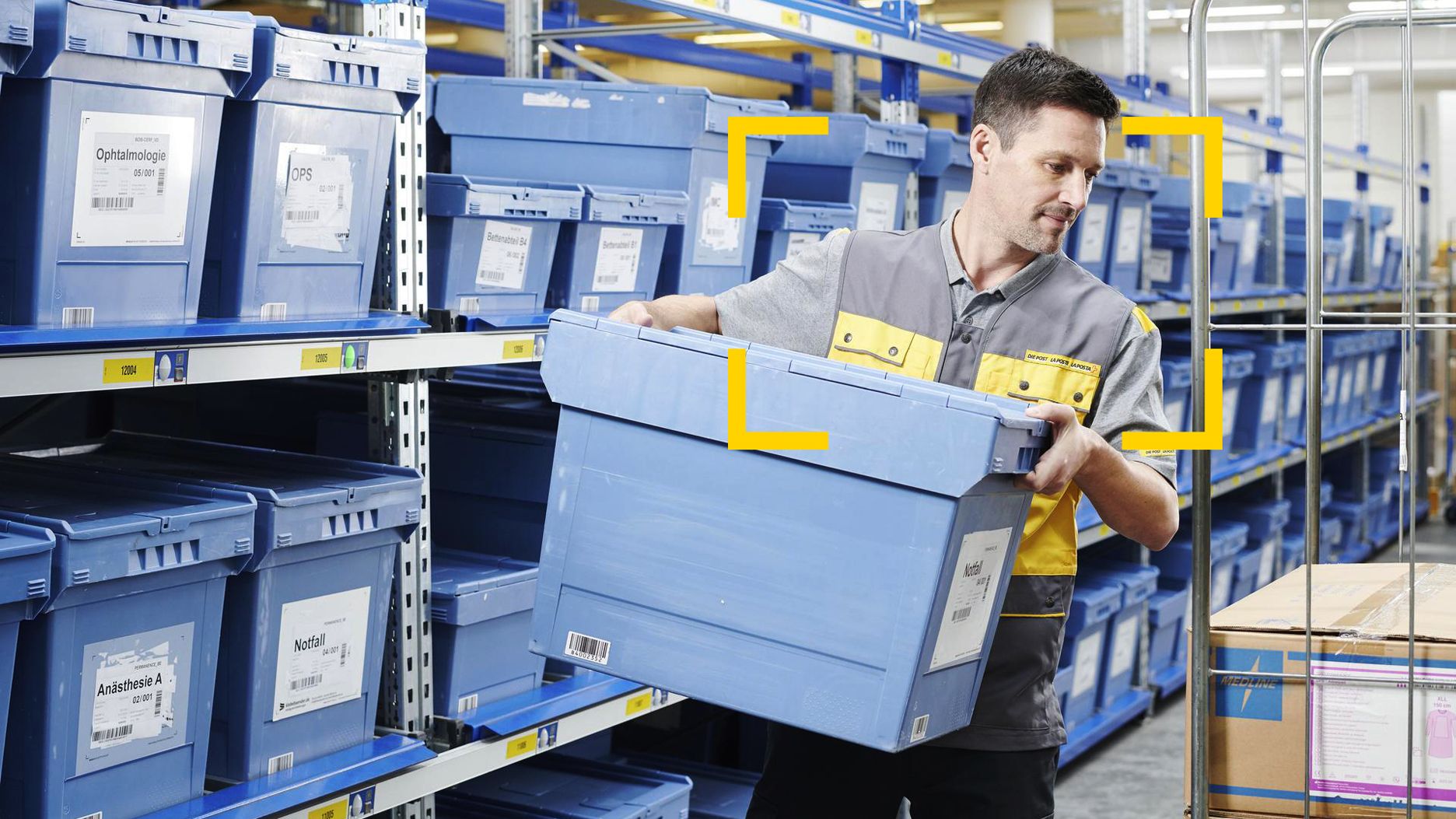 Eine Person hebt in einem Lagerraum eine grosse blaue Kunststoffbox aus einem Regal. Die Umgebung besteht aus mehreren Reihen gleichartiger blauer Boxen, die in Metallregalen gestapelt sind. Die Person trägt Arbeitskleidung in Grau und Gelb.