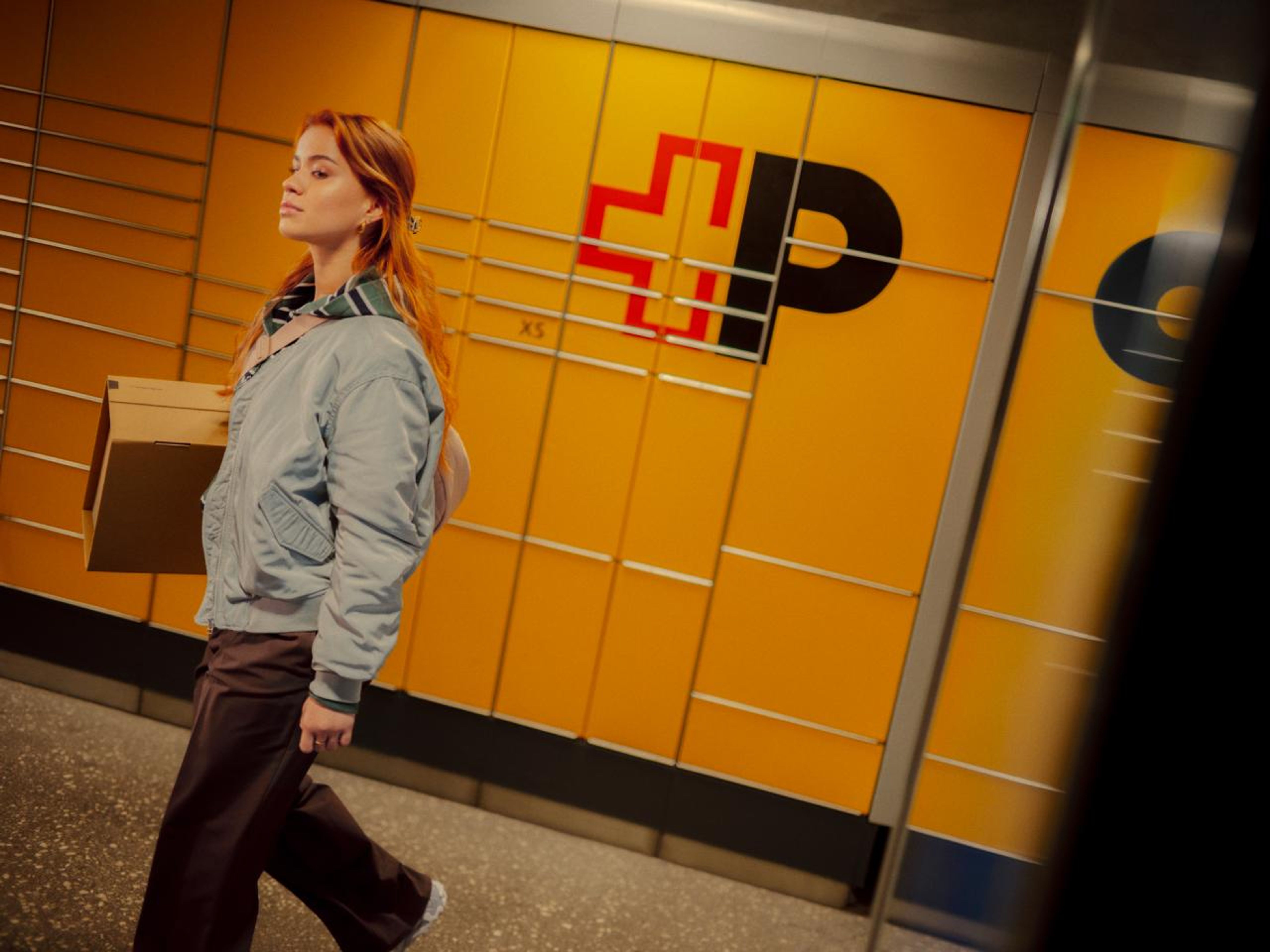 Eine Person trägt ein Paket und läuft an einer gelben Postfachanlage "My Post 24" vorbei. Die Anlage ist mit dem Logo der Schweizerischen Post versehen und befindet sich in einem modernen, sauberen Innenraum. Die Person wirkt entspannt und der Raum ist gut beleuchtet.