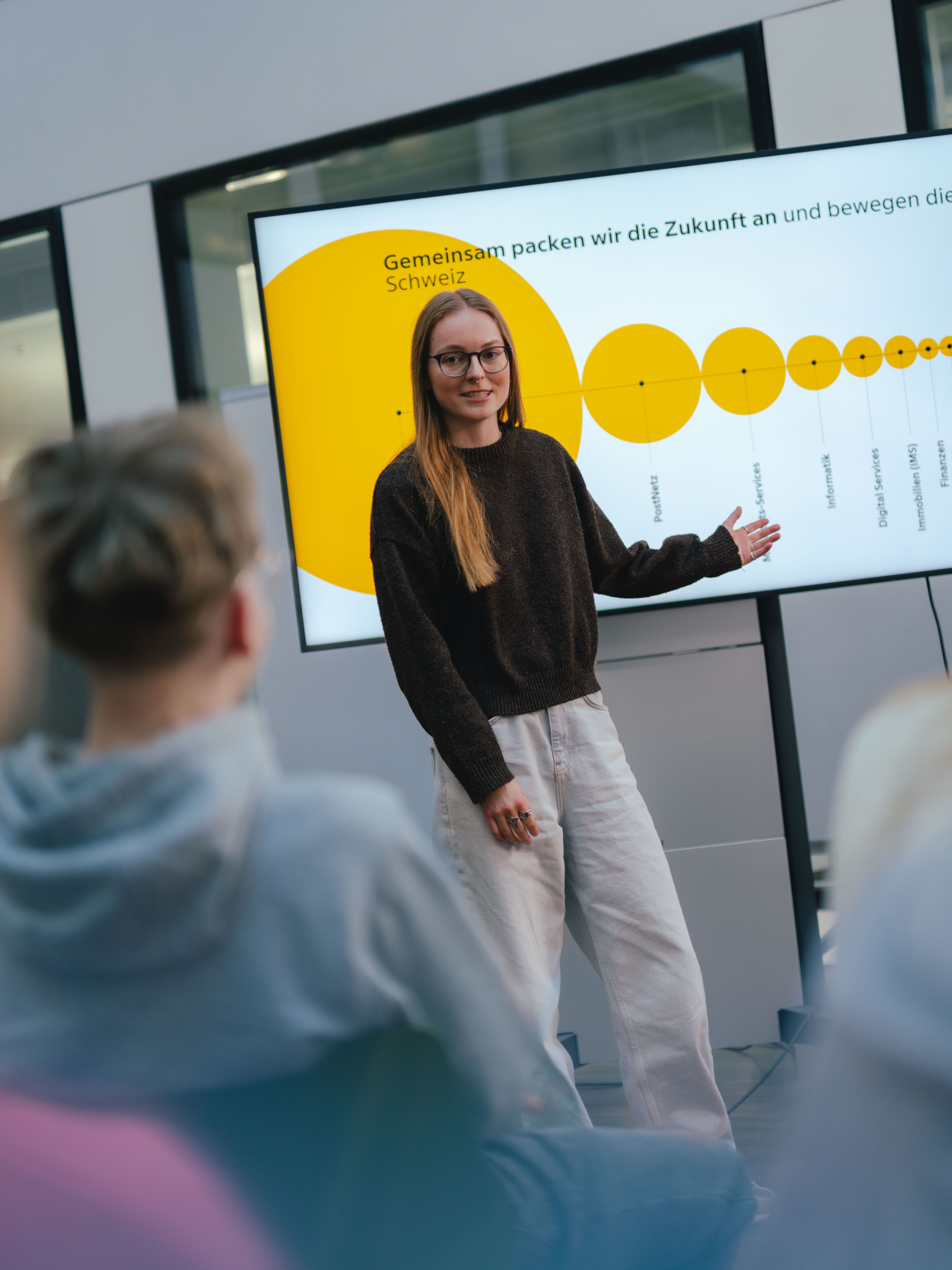 Eine Person steht vor einem grossen Bildschirm. Auf dem Bildschirm ist eine gelbe Grafik mit unterschiedlich grossen Kreisen zu sehen, verbunden in einer Linie. Vor der stehenden Person sitzen mehrere weitere Personen und schauen in Richtung Präsentation. Der Raum wirkt wie ein moderner Besprechungs‑ oder Schulungsraum.