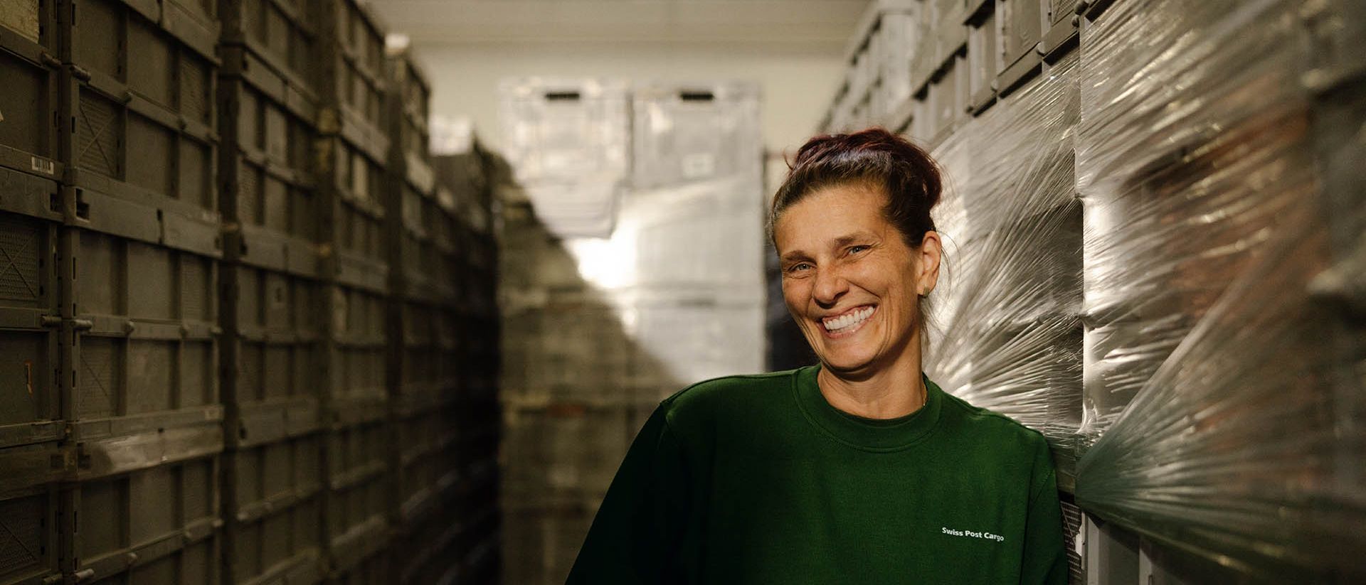 A logistics employee from Swiss Post Cargo is smiling while standing between stacked transport crates in a warehouse.