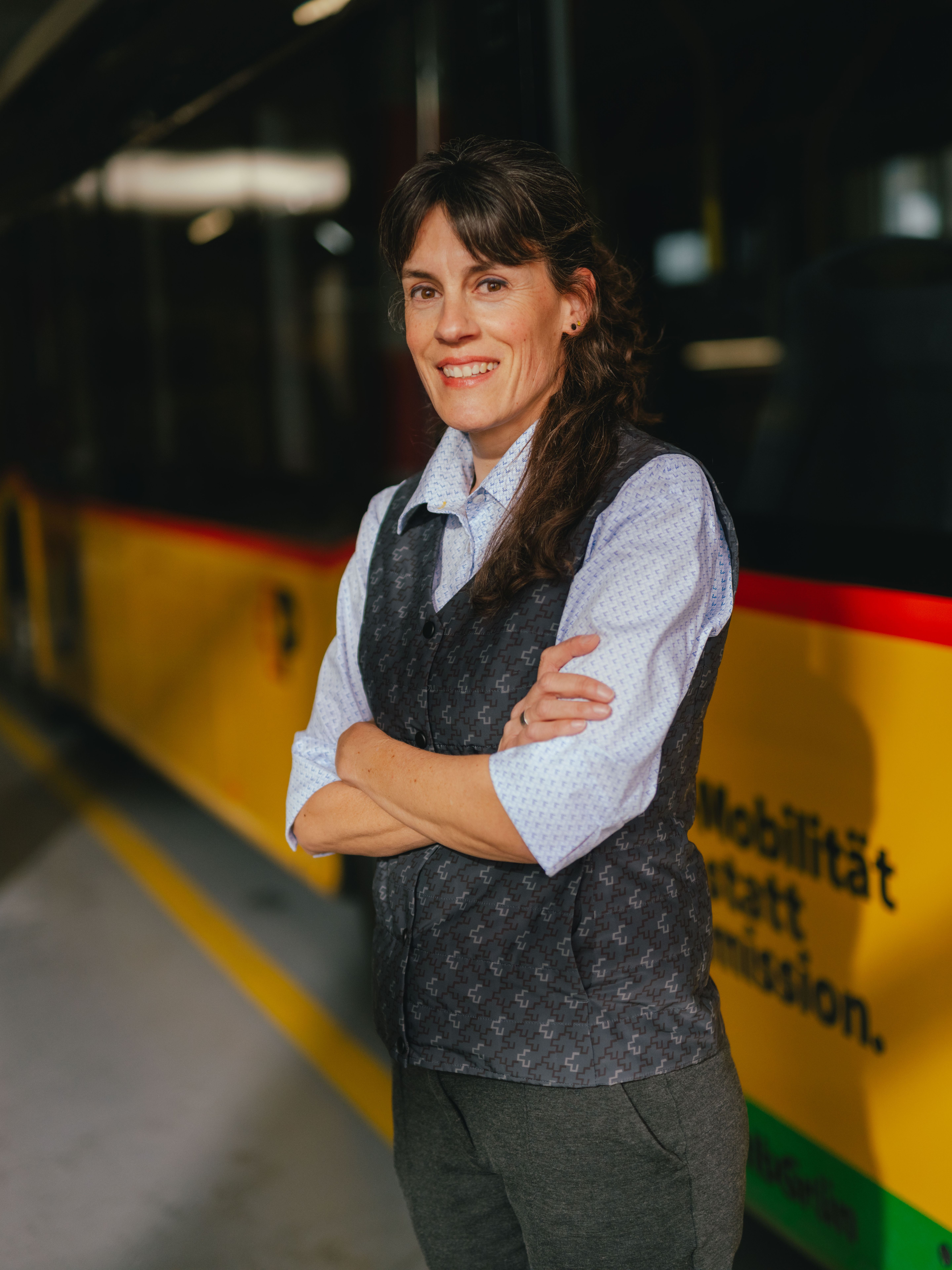 Una persona è in piedi con le braccia incrociate in un capannone vicino a un veicolo giallo e nero. La persona indossa una camicia chiara e un gilet scuro. L’ambiente sembra essere un deposito o un’area di parcheggio, con colonne a strisce e pavimento riflettente.